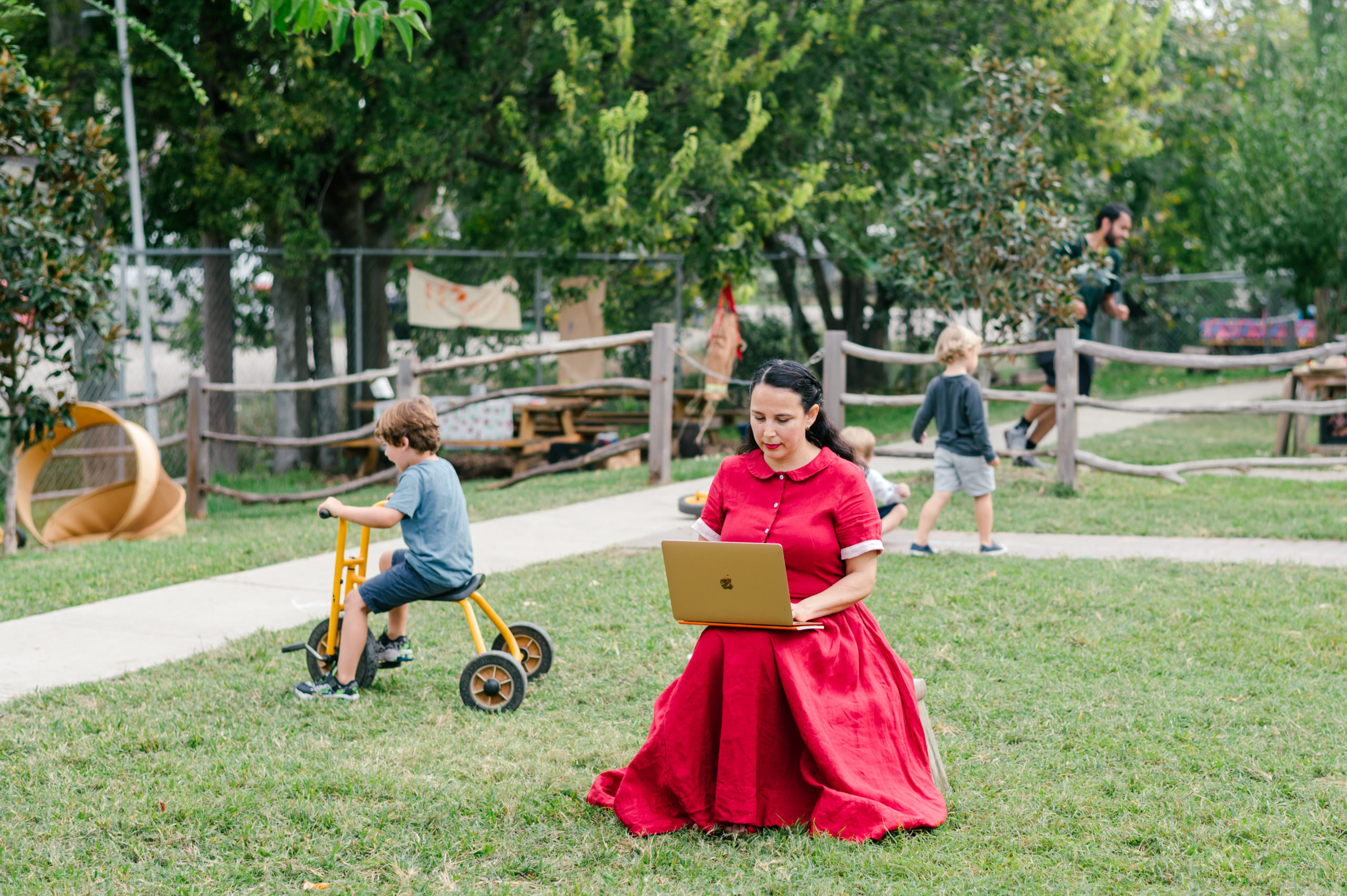 Photo of woman child development coach in a beautiful red dress observing kids on the playground while taking notes on her computer  during her branding photography session
