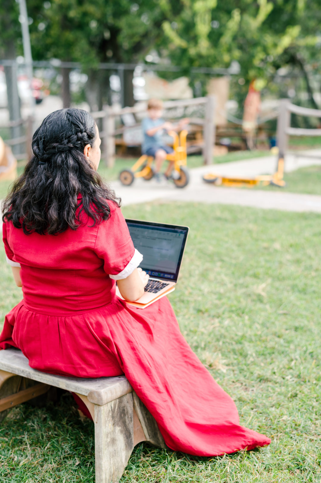 Photo of woman child development coach in a beautiful red dress observing kids on the playground while taking notes on her computer  during her branding photography session
