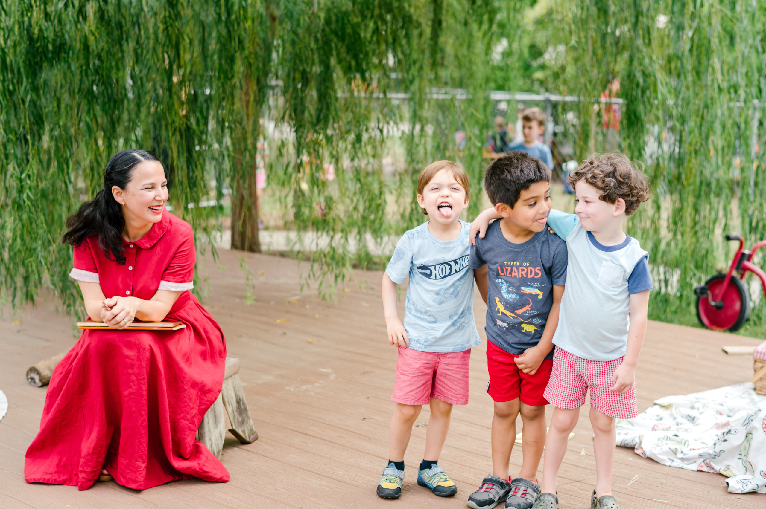 Photo of woman child development coach in a beautiful red dress smiling at kids on a playground while they smile and stick their tongues at to the camera 