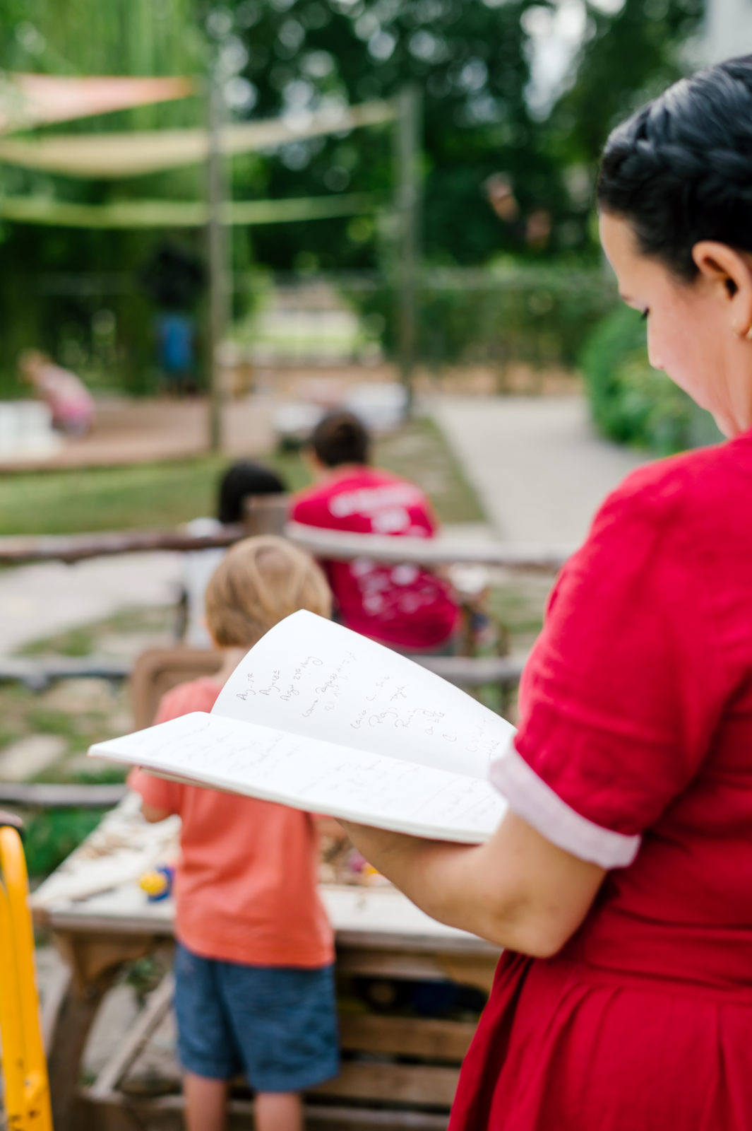 Photo of woman child development coach in a beautiful red dress observing kids on the playground while taking notes in her notebook  during her branding photography session
