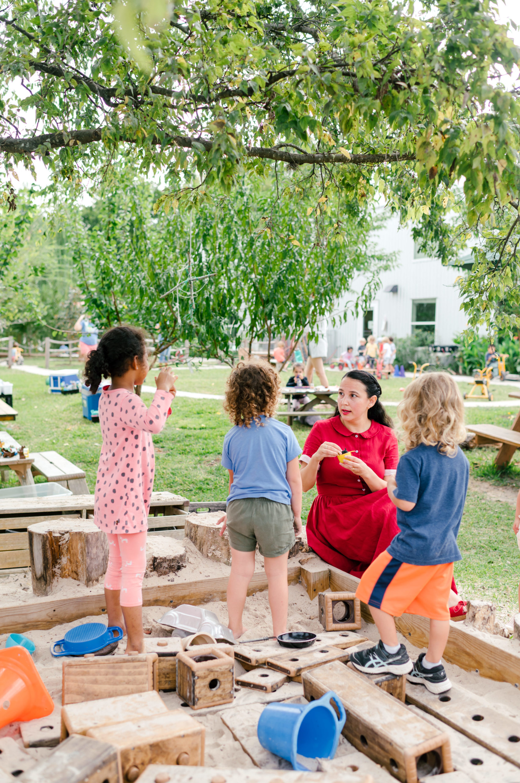 Photo of woman child development coach in a beautiful red dress interacting with kids on a playground outside in a sandbox during her branding photography session