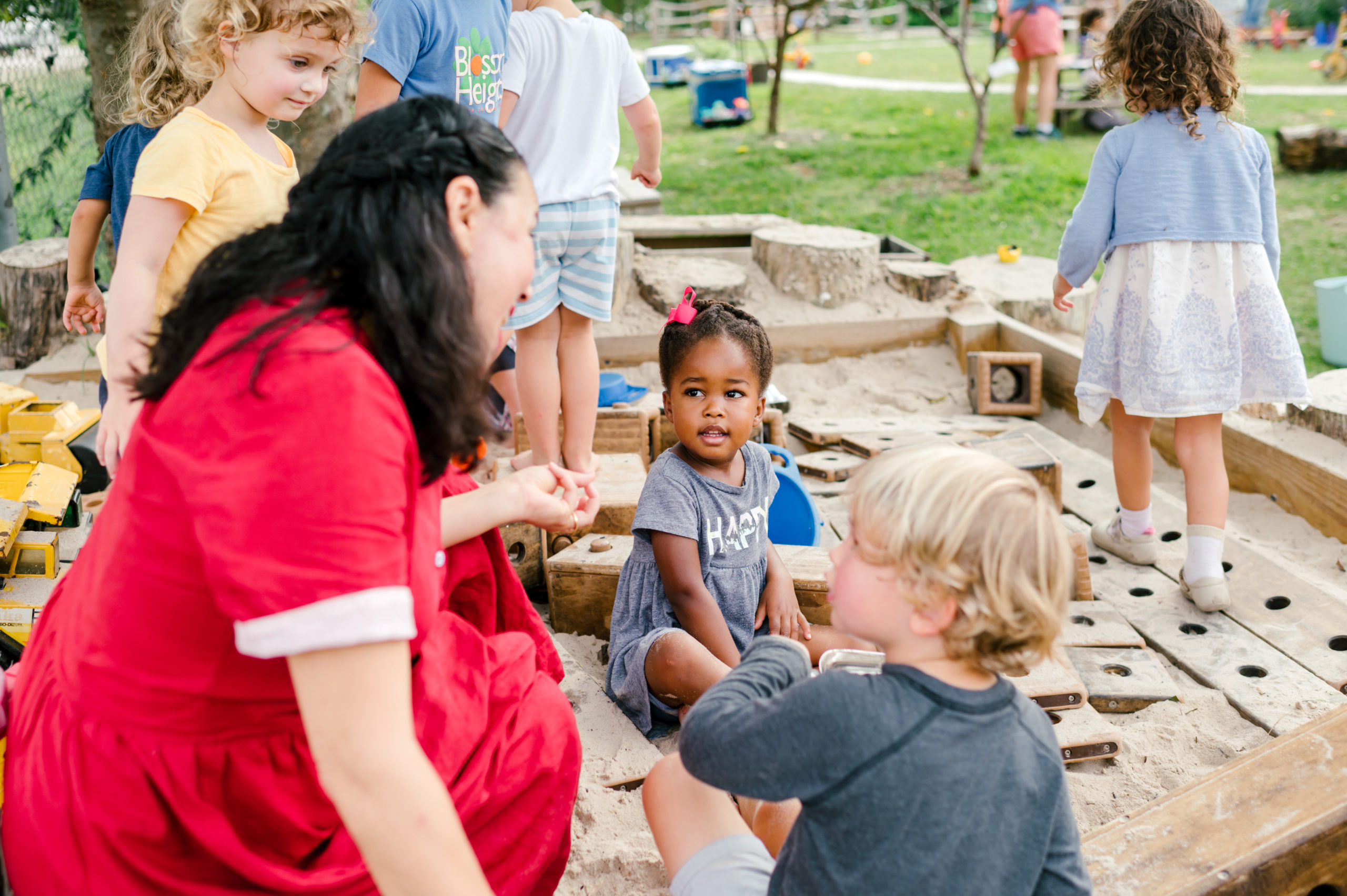 Photo of woman child development coach in a beautiful red dress interacting with kids on a playground outside in a sandbox during her branding photography session