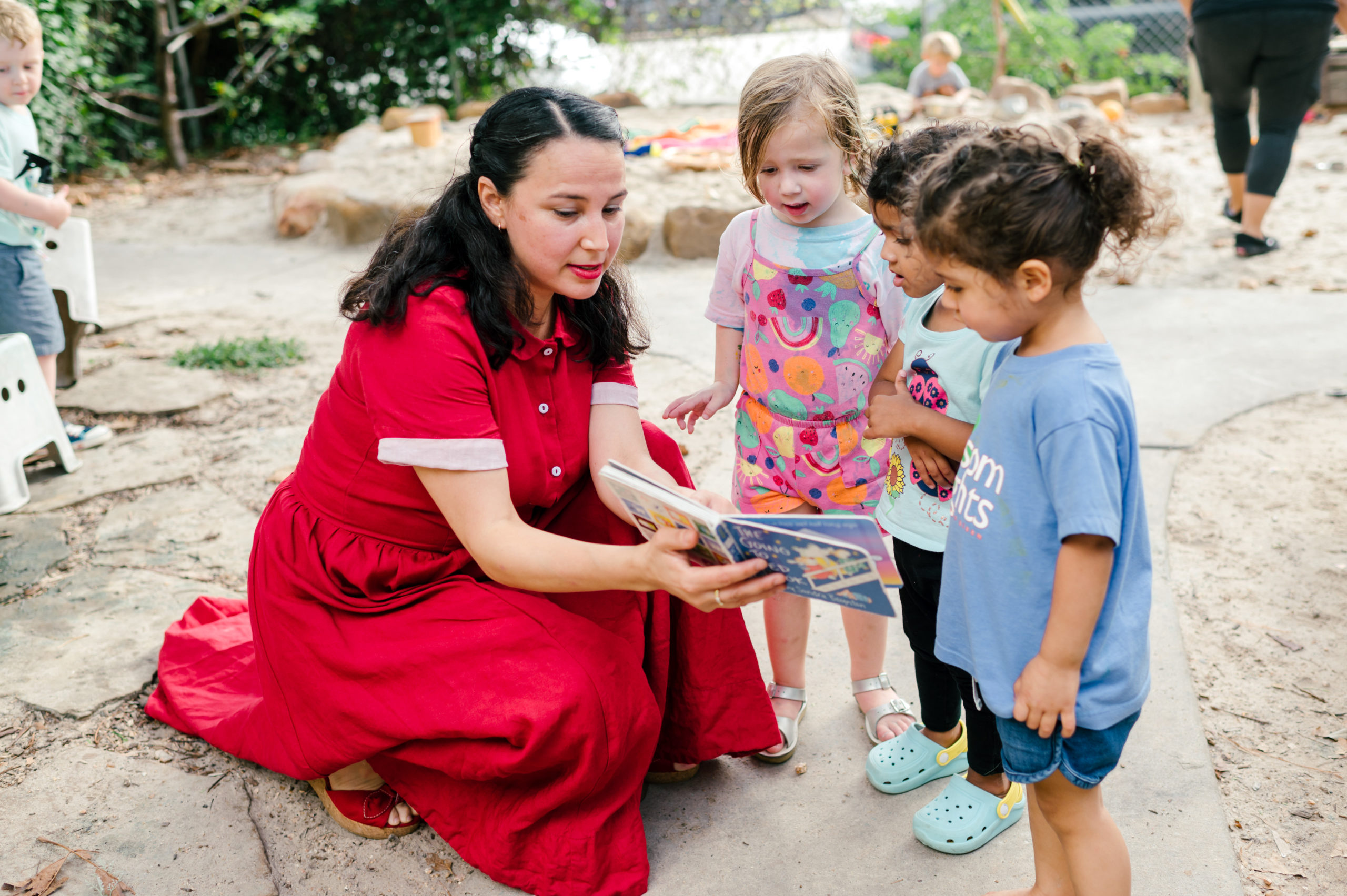 Photo of woman child development coach in a beautiful red dress interacting with kids on a playground outside reading a childs book during her branding photography session