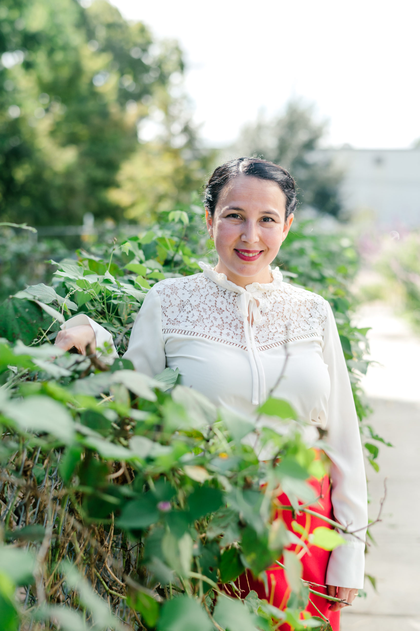 Photo of woman child development coach standing next to a bush in a white button up and red pink skirt and red heels smiling 
