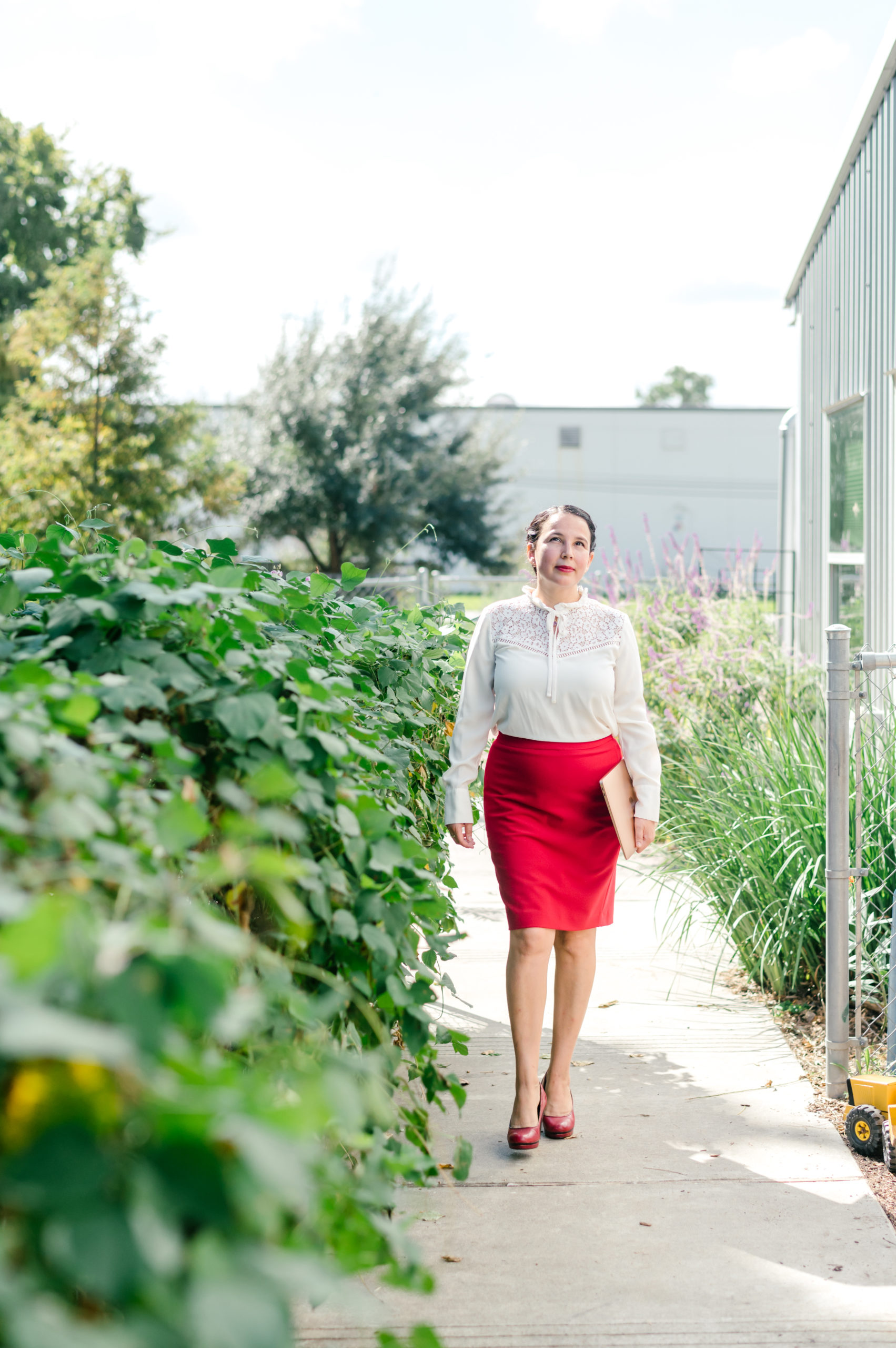 Photo of woman child development coach standing next to a bush in a white button up and red pink skirt and red heels 

