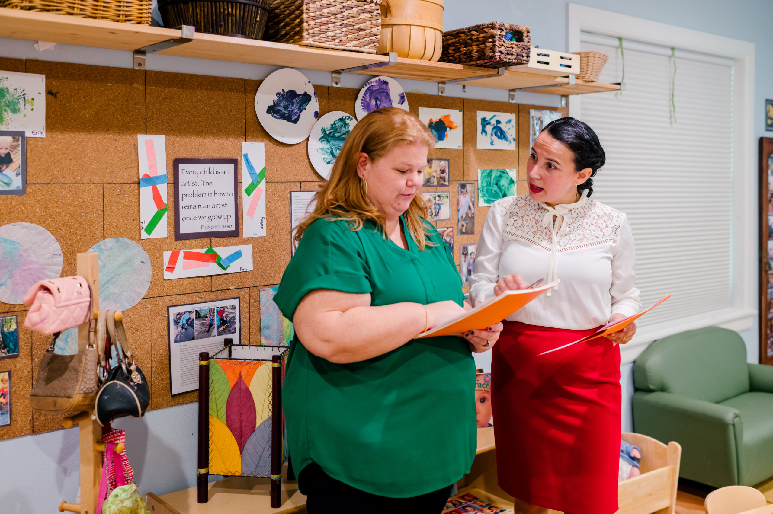 Photo of woman child development coach in a white button up and red pink skirt speaking to a teacher in a school classroom 