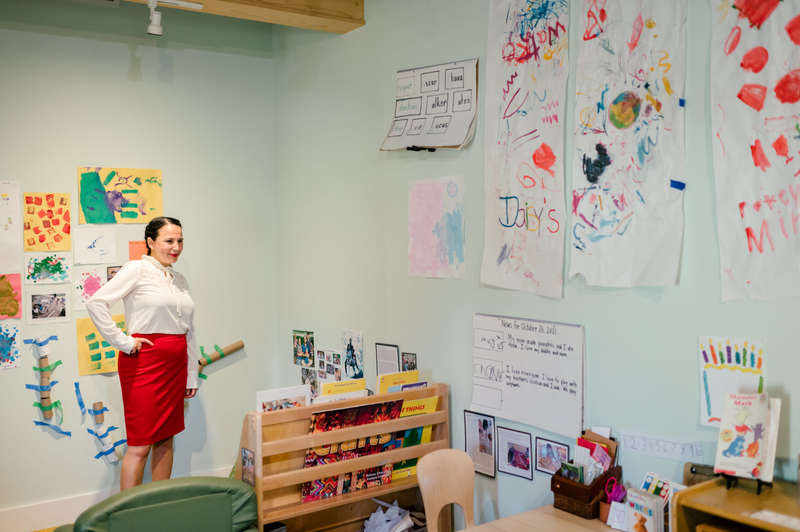 Photo of woman child development coach standing smiling in a white button up and red pink skirt and red heels observing kids in a school classroom
