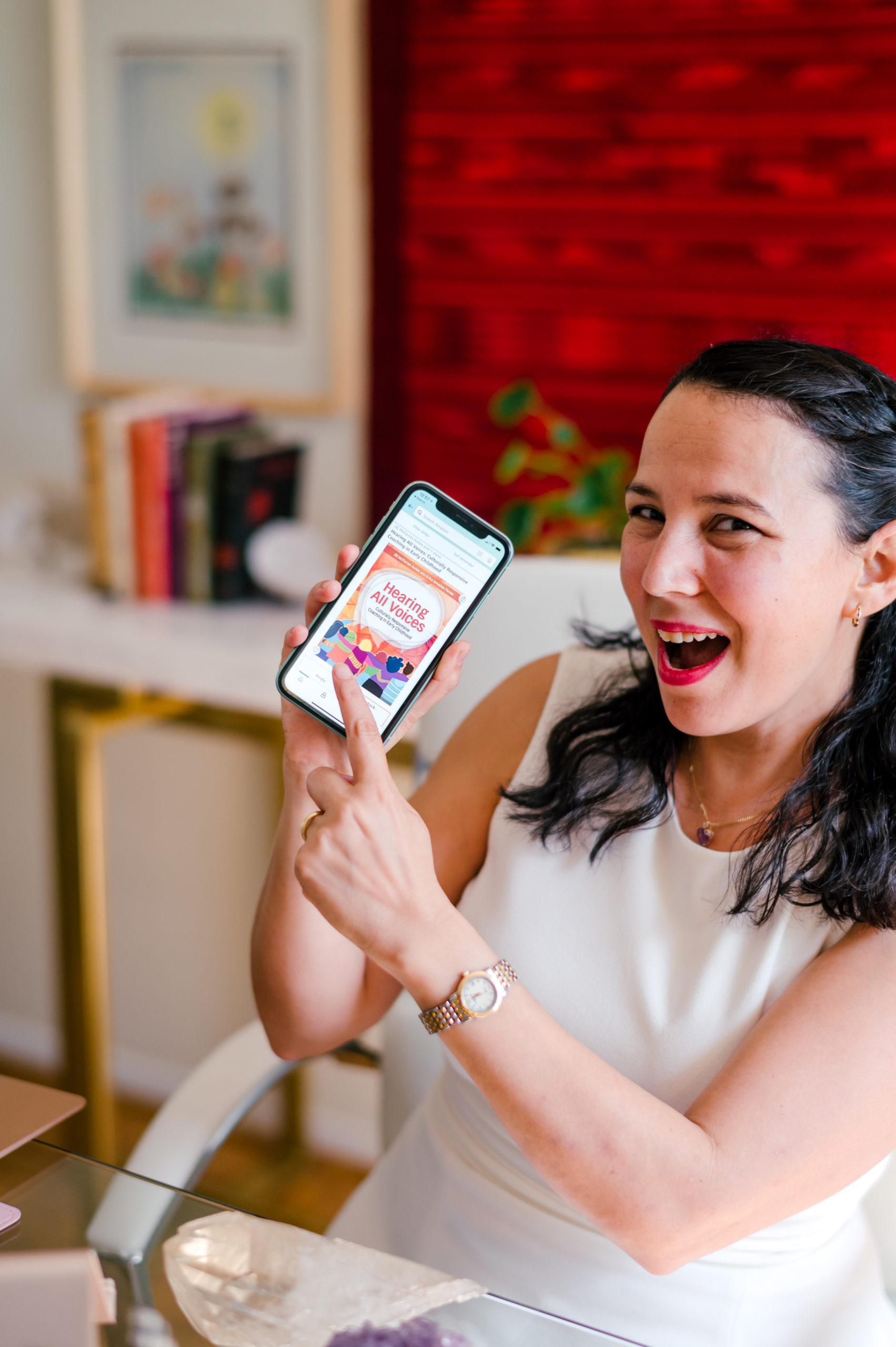 Photo of woman child development coach in a white dress pointing to her iPhone with a picture of the front cover of the book Hearing All Voices  during her branding photography session