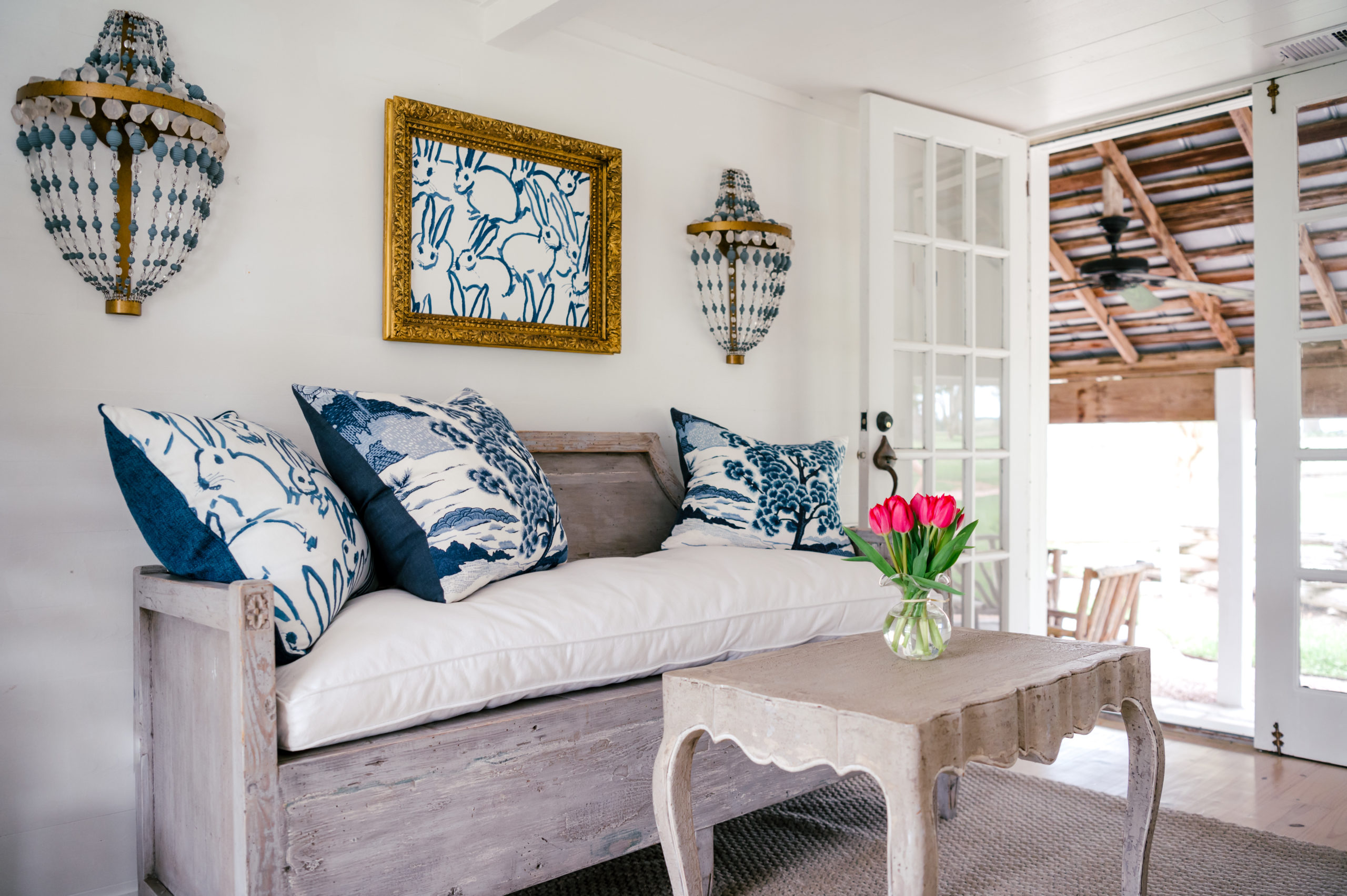 Interior Photography of wooden couch and white and blue cushions in a room with a frame of blue bunnies and beautiful chandler lamps and a grey table with pink flowers on the table