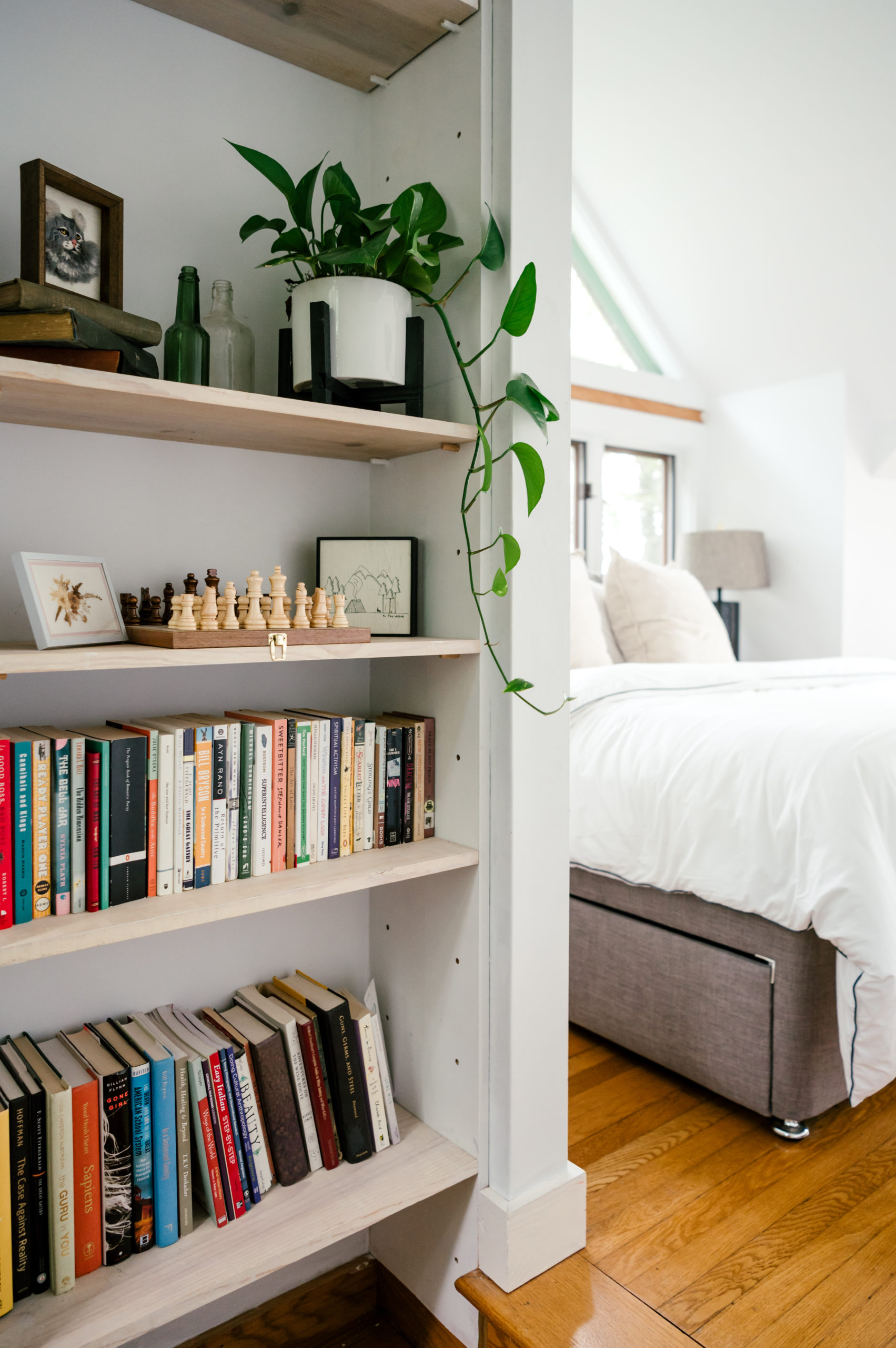 Photo of bedroom interior decor such as books and a chess board sitting on the shelves, and bed with white cover in the background