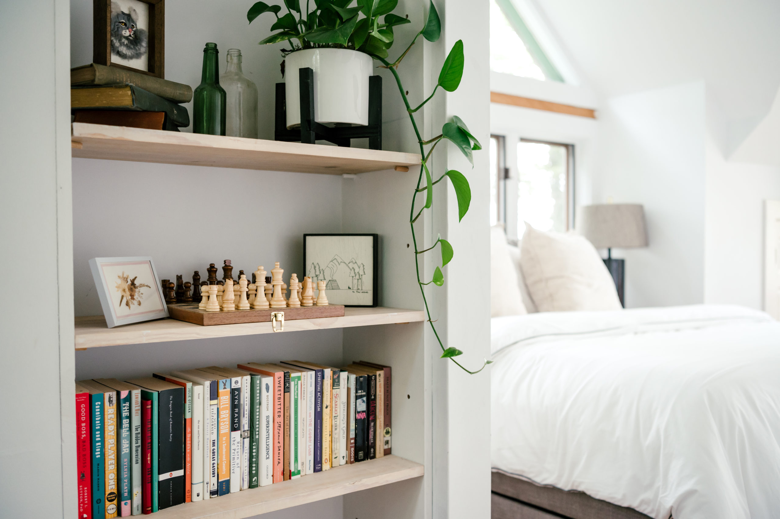 Photo of bedroom interior decor such as books and a chess board sitting on the shelves, and bed with white cover in the background