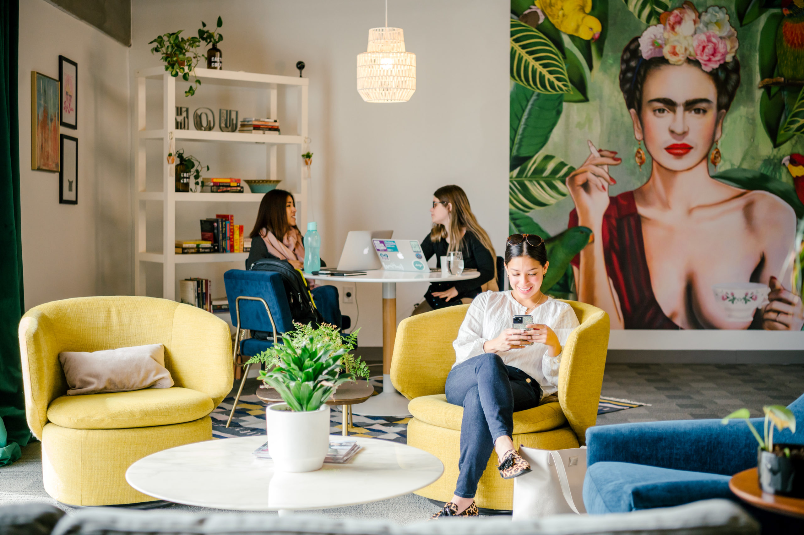 Photo of office interior with people sitting in lounge chairs on their phones and Frida Kahlo art on the wall in the background 