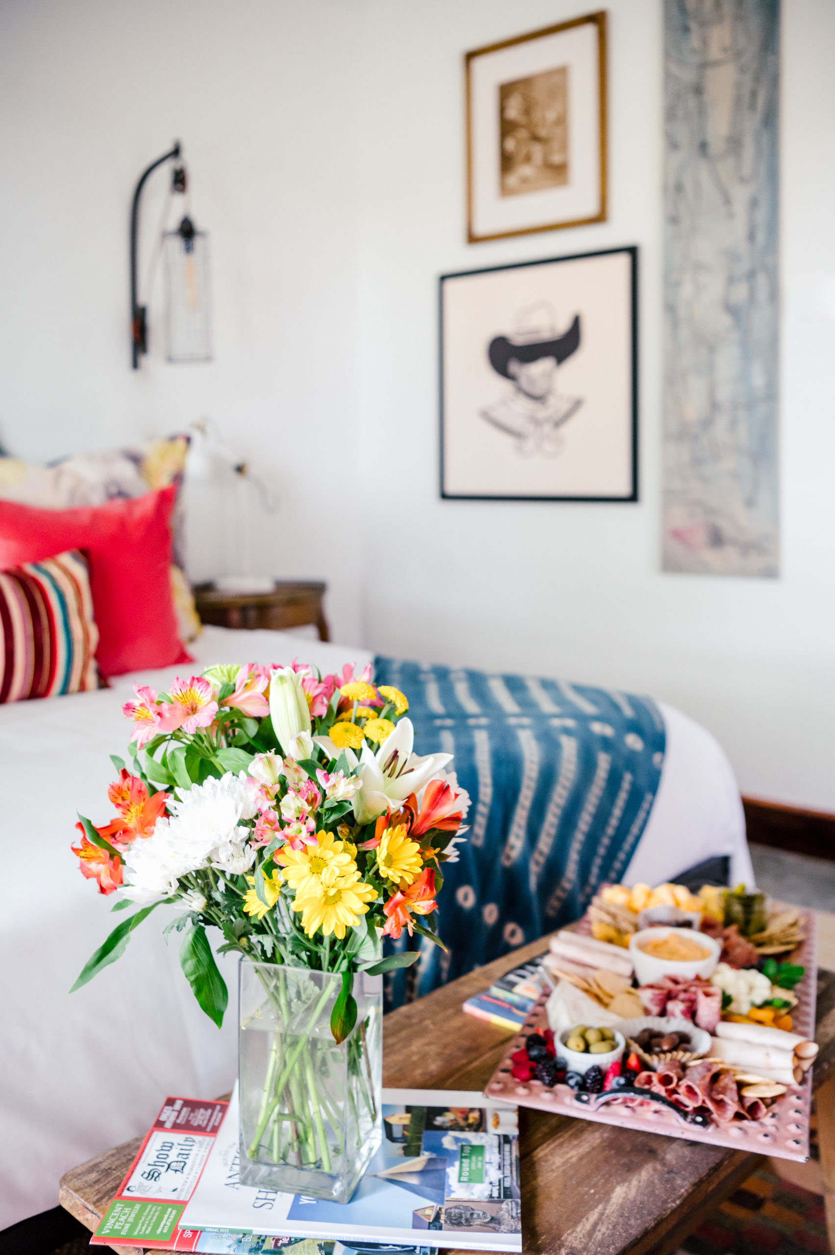Room with a queen sized bed with colorful pillows and western art hanging on the wall with a bench at the end of the bed with flowers and a charcuterie board 