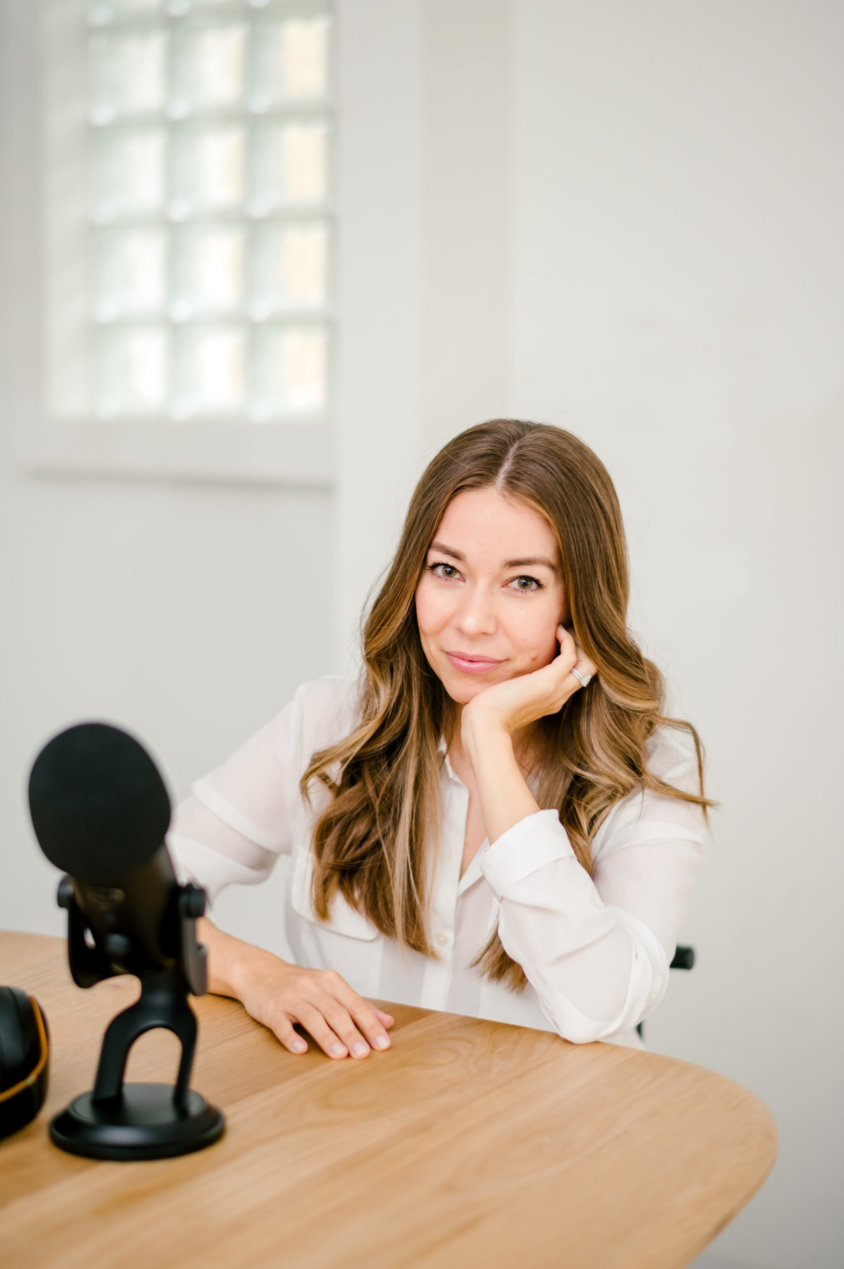 Woman sitting leaning on a wooden table perched on her cheek next to podcast mic for her brand photography photos