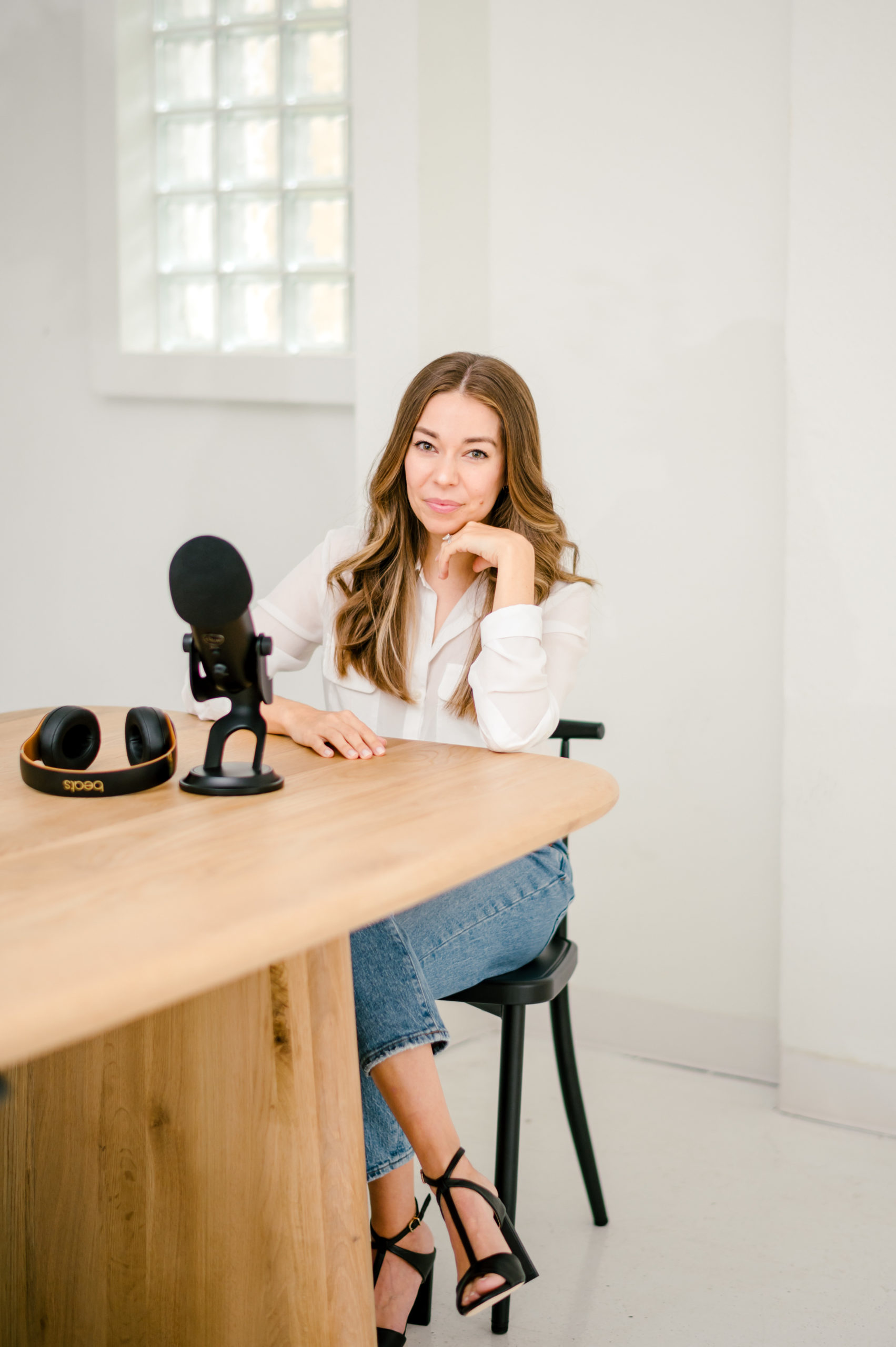 Woman in a white button up and jeans sitting down leaning on wooden table next to a podcast mic for her brand photography photos