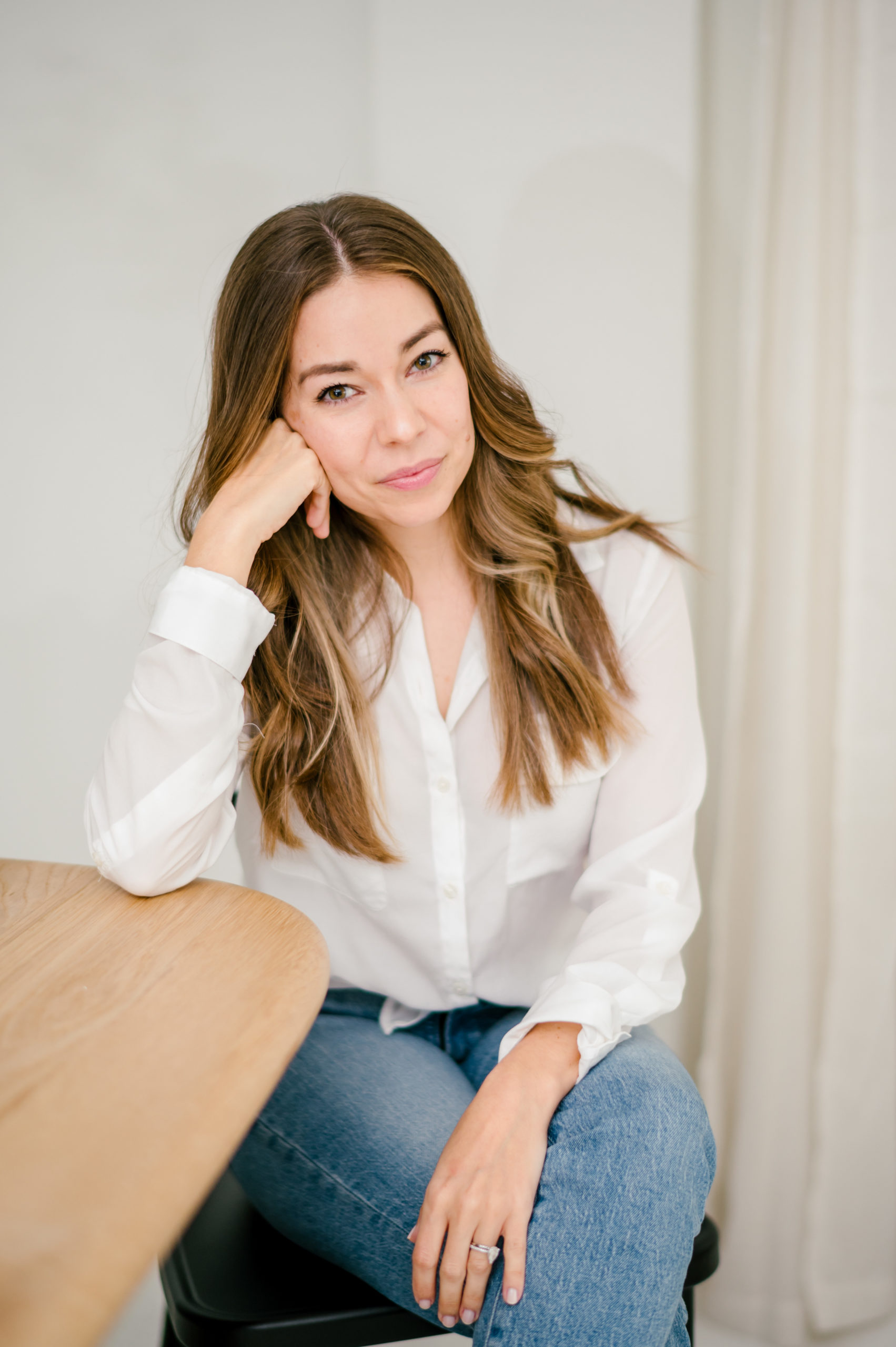 Woman in a white button down and jeans sitting leaning on a wood table with her hand resting on her face for her brand photography photos