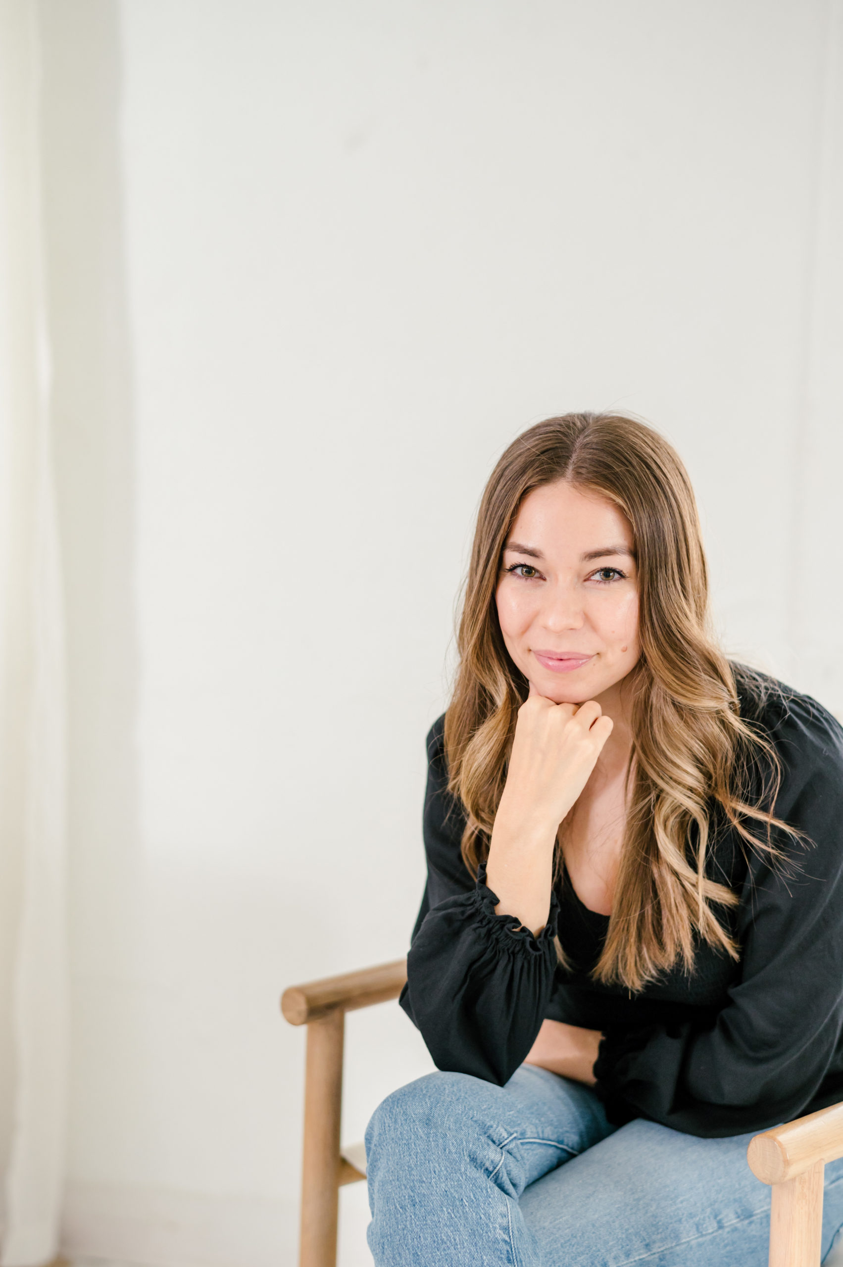 Woman sitting on on wooden bench leaning on her knees smiling in a black blouse and jeans 