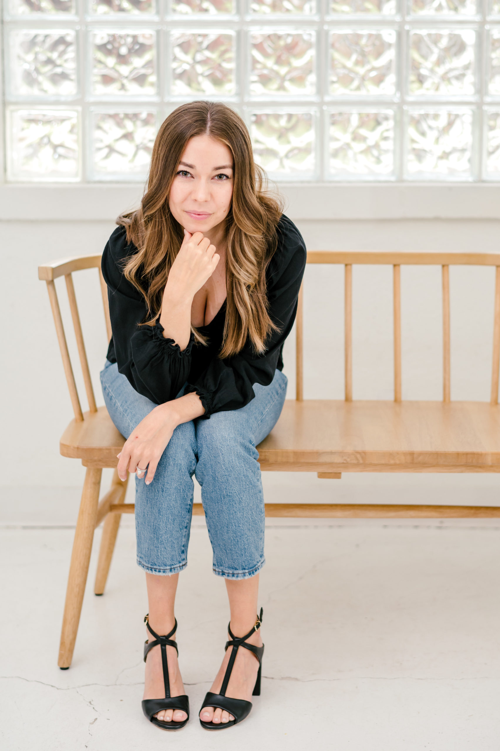 Woman sitting on on wooden bench leaning on her knees smiling in a black blouse and jeans 