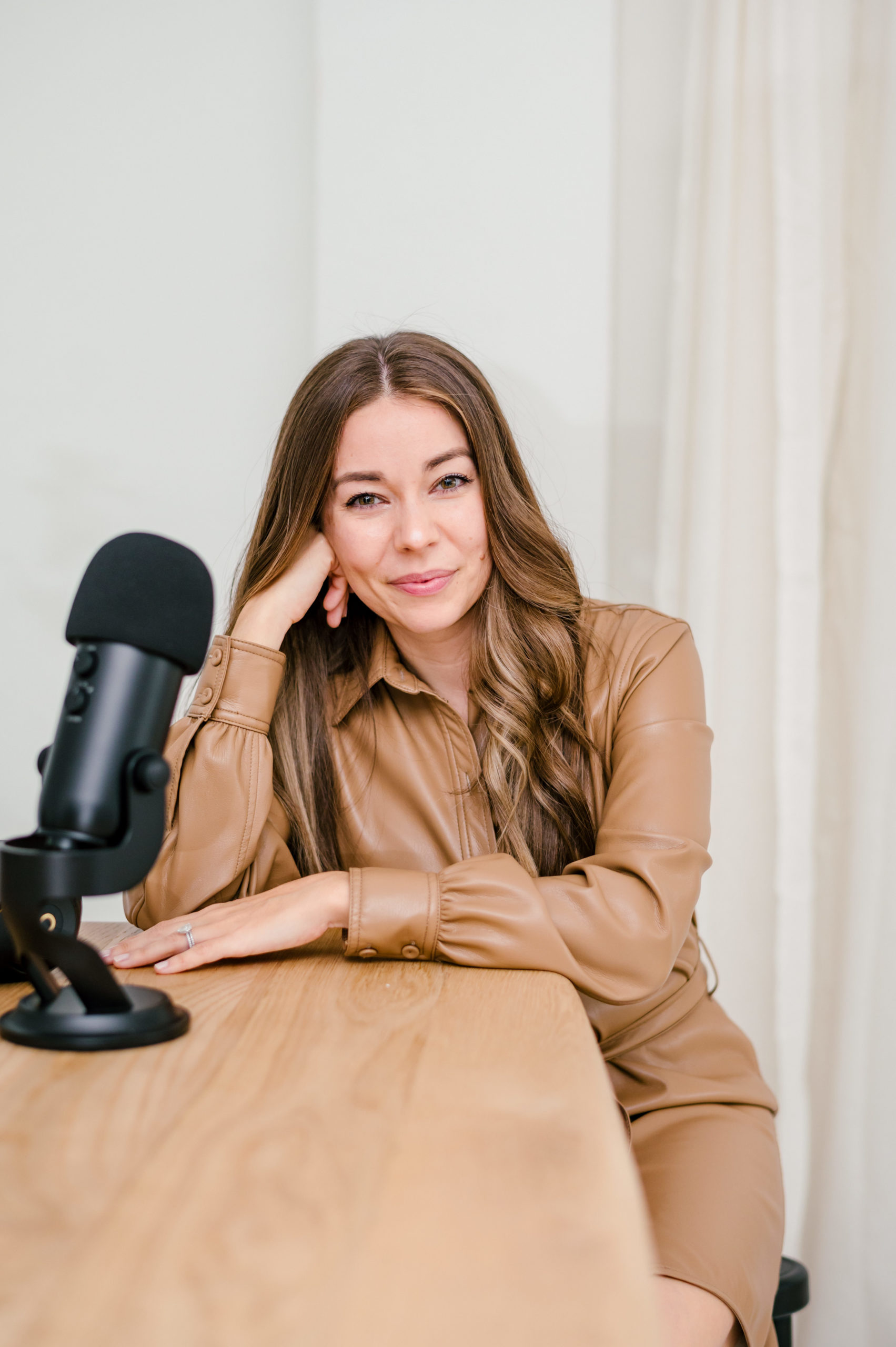 Woman in a leather dress leaning on wooden table next to a podcast mic