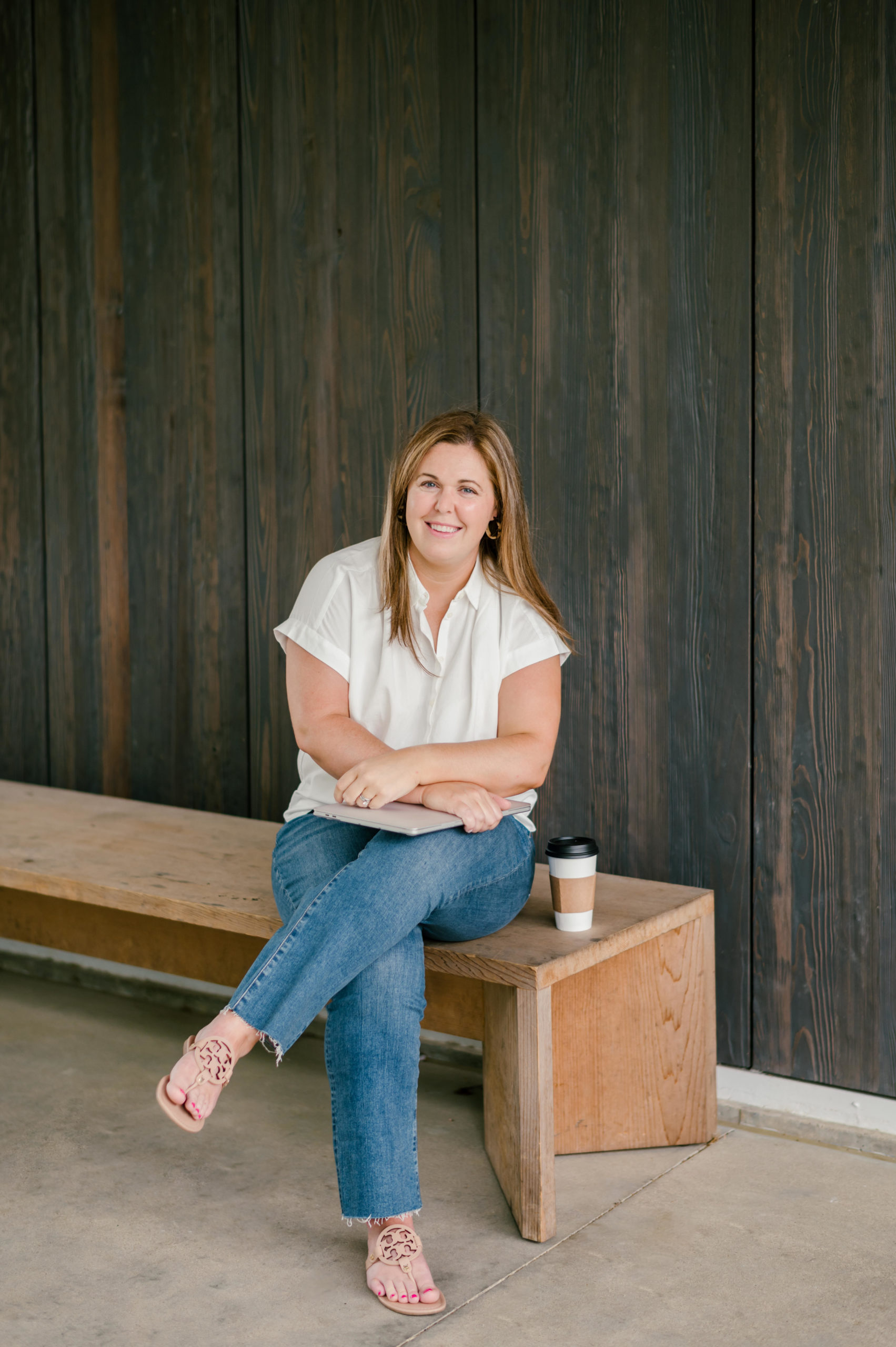 Woman sitting on a wooden bench criss crossing and smiling in a white blouse and blue jeans her personal brand photos