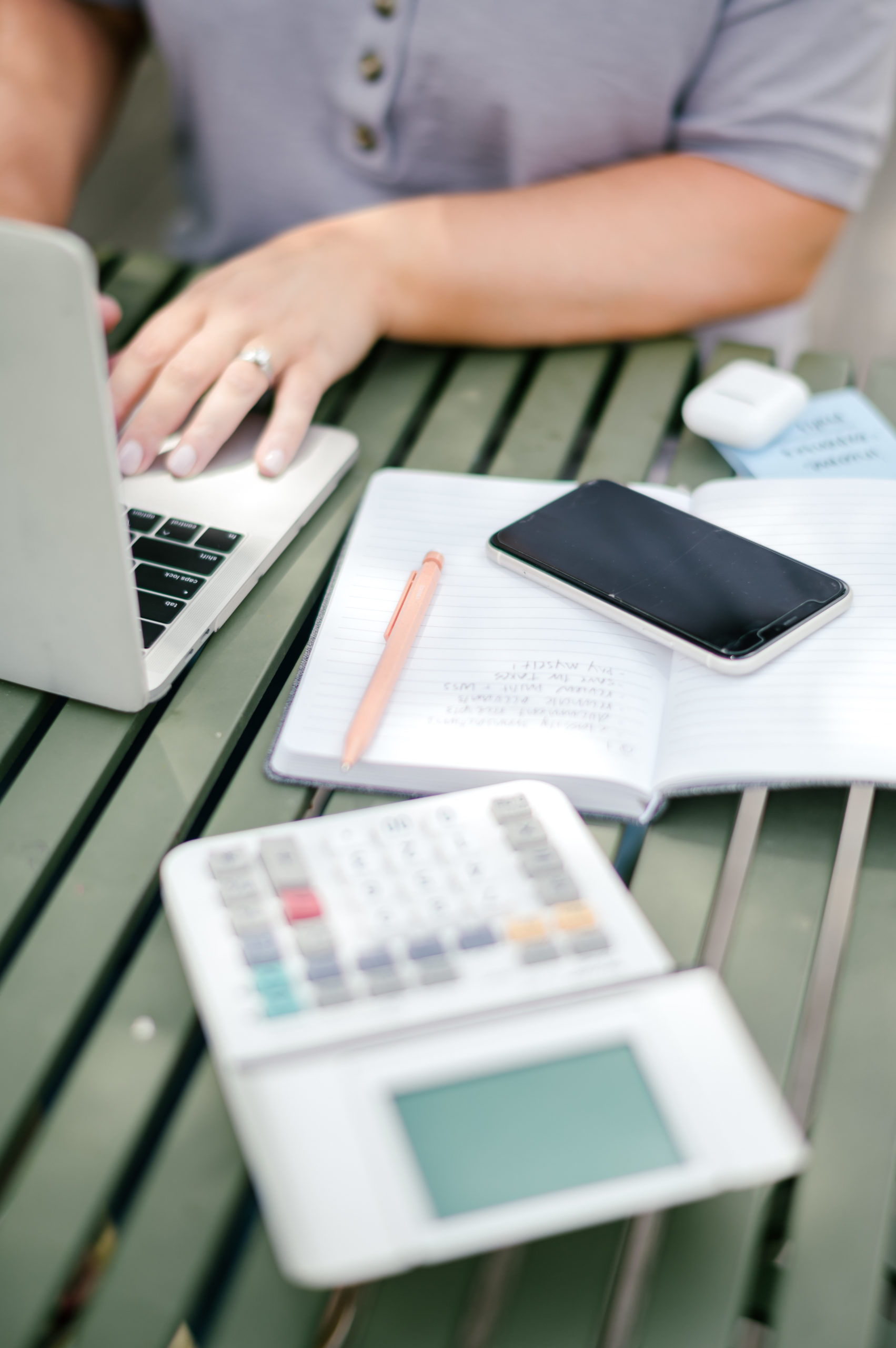 Woman accountant working on a green bench outside typing on her laptop with a notebook, calculator and phone on her side