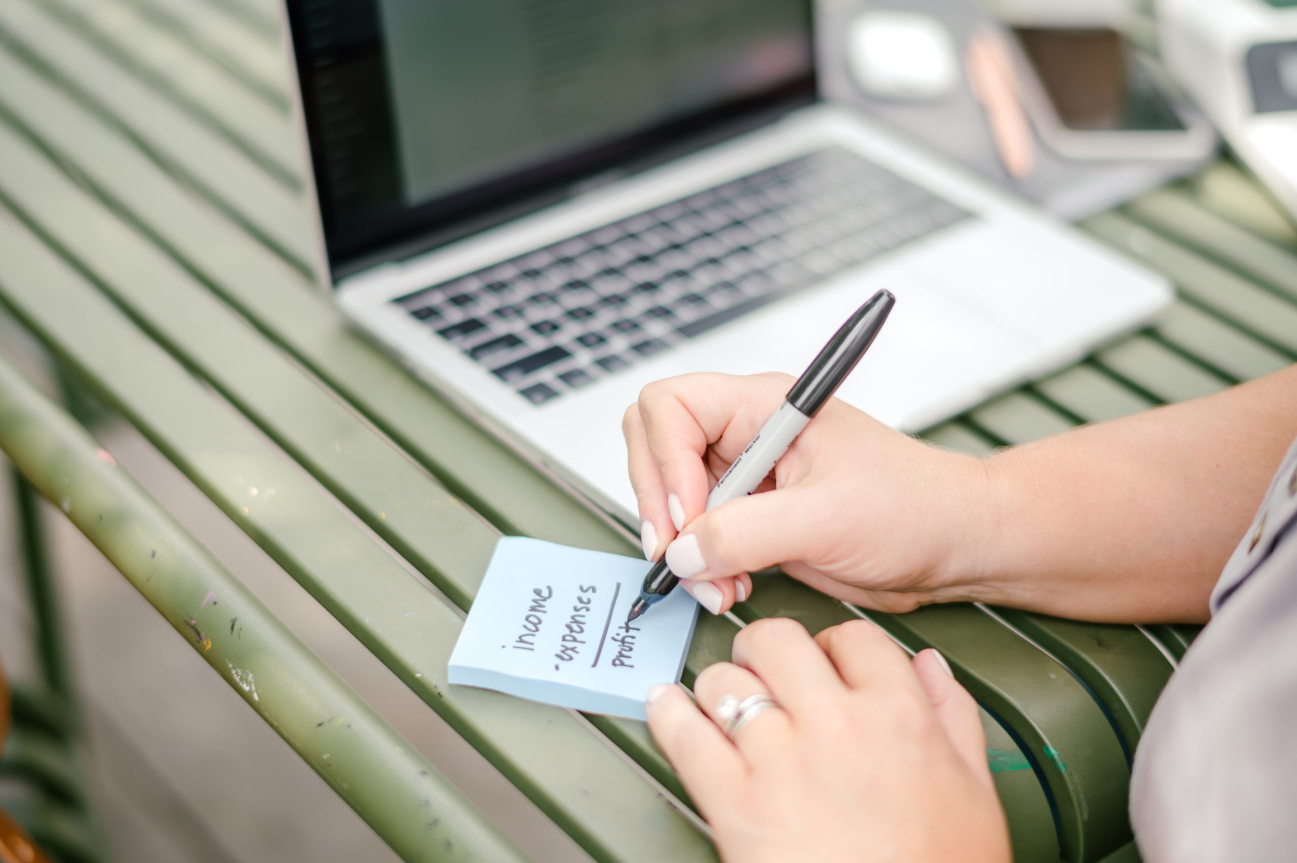 Woman accountant writing notes on a blue notepad while working on her laptop on a green outside table