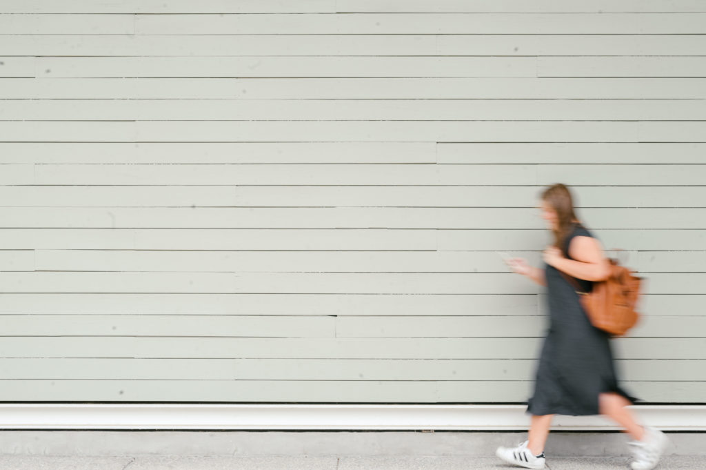 Woman walking down the side of the street in a grey t-shirt dress holding a brown leather backpack 