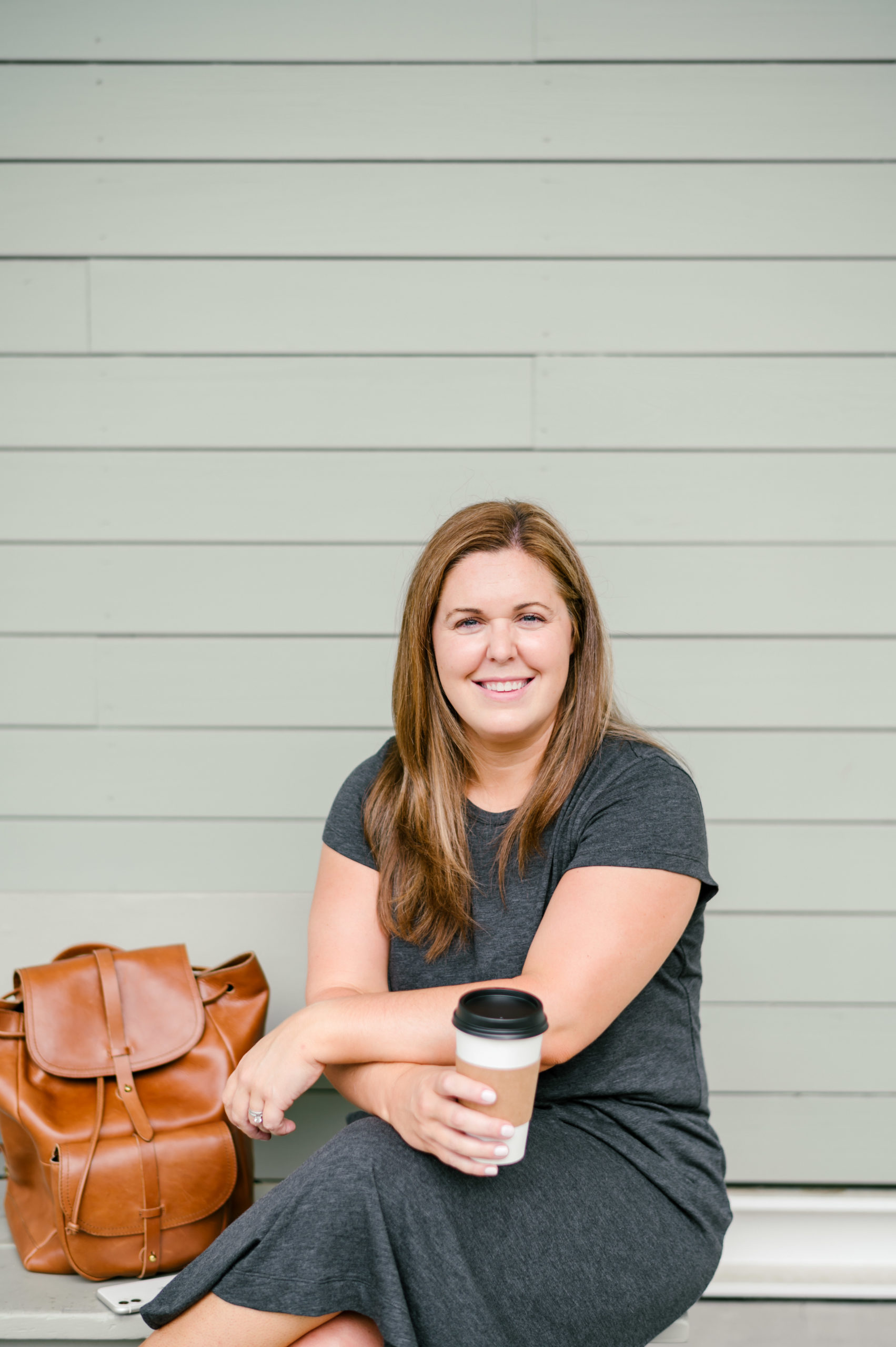 Woman sitting on a metal bench holding a cup of coffee smiling in a grey t-shirt dress her personal brand photos