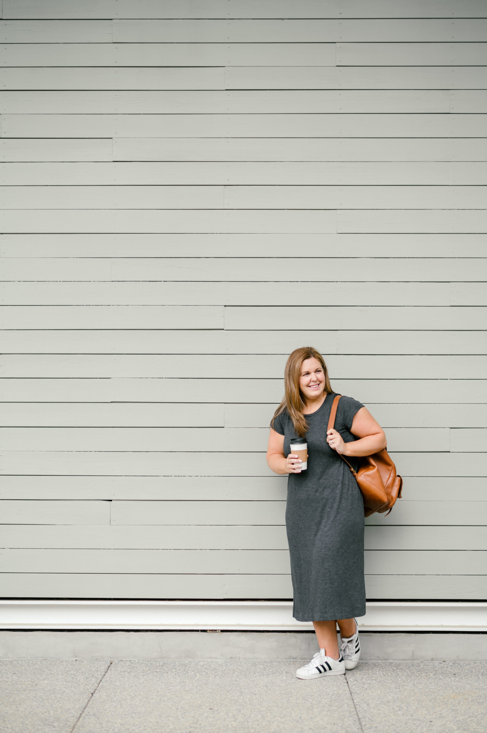 Woman standing holding a cup of coffee in a grey t-shirt dress holding a brown leather backpack her personal brand photos