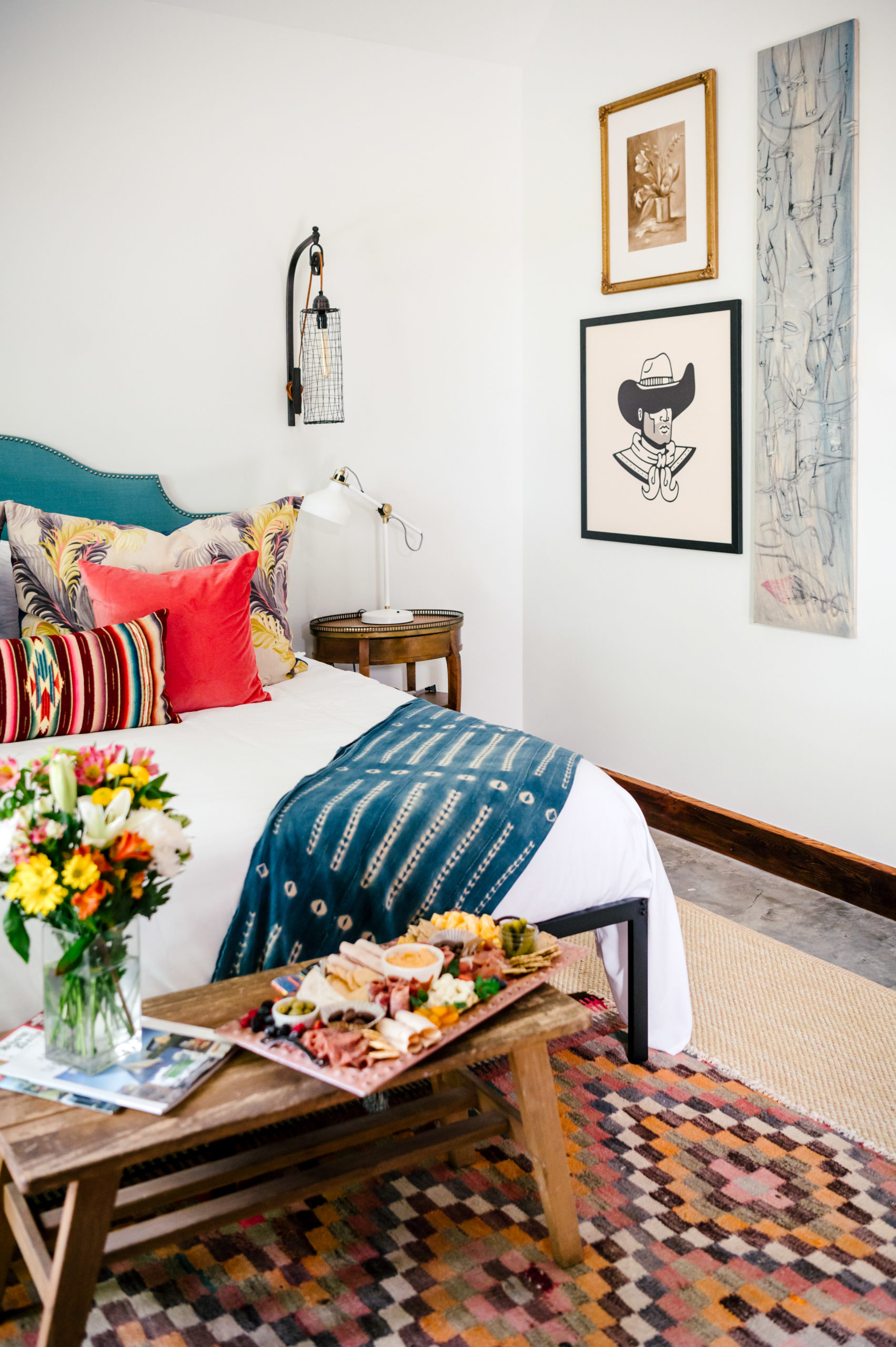 Bedroom interior with a bed filled with colorful pillows and country art hanging on the wall and a Charcuterie board sitting at the end of the bed on a coffee table 