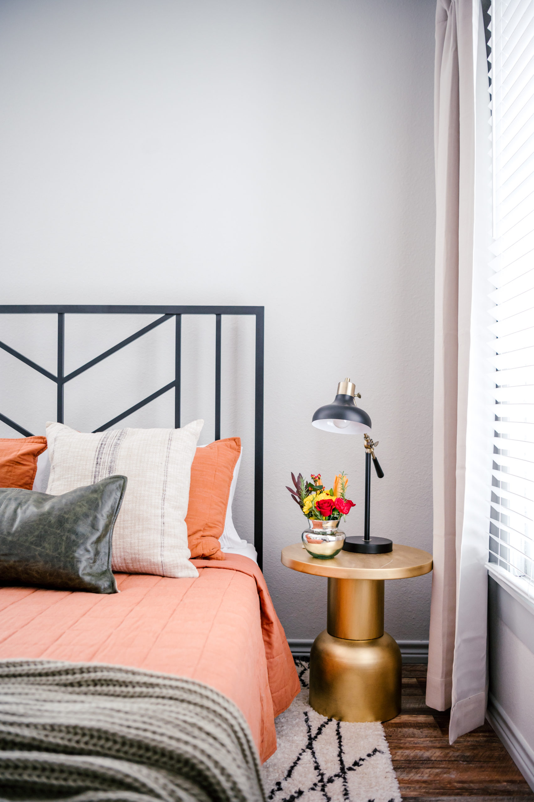 Bedroom interior with queen bed made with an orange comforter and pillows, and gold side table with a black lamp