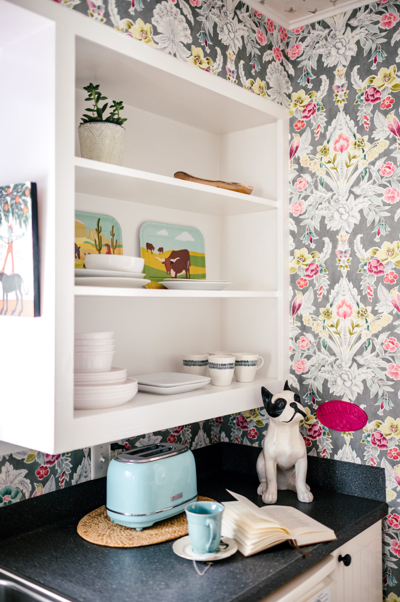 Short-term Rental Photos of kitchen interior with colorful wallpaper, grey countertops, and dishware on shelving