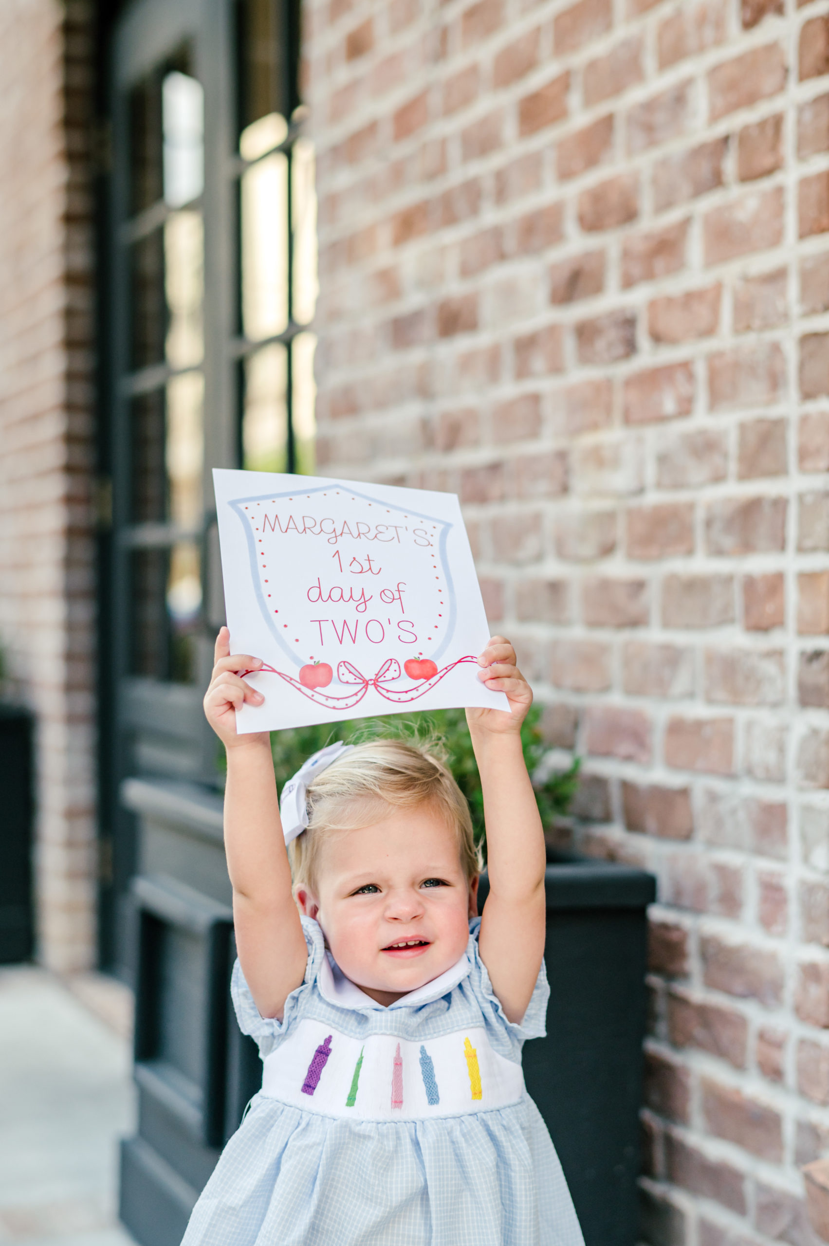 Little girl holding a milestone card labeled "Margaret's 1st day of two's 