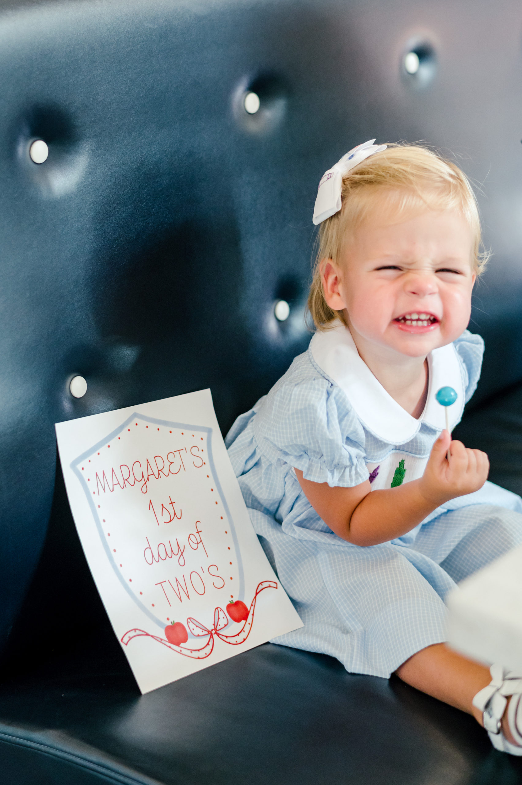 Baby smiling holding a lollipop sitting next to milestone card labeled Margarets 1st day of two's