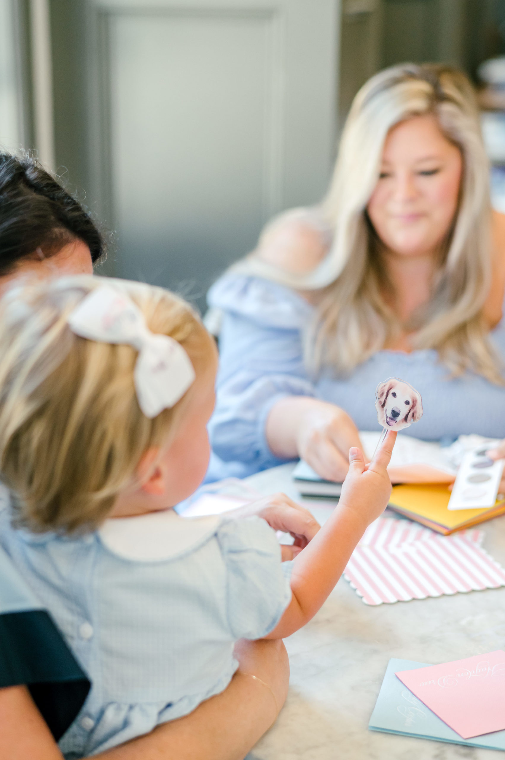 Baby holding a puppy sticker on finger while Woman makes a paper invitation for her etsy shop in the background