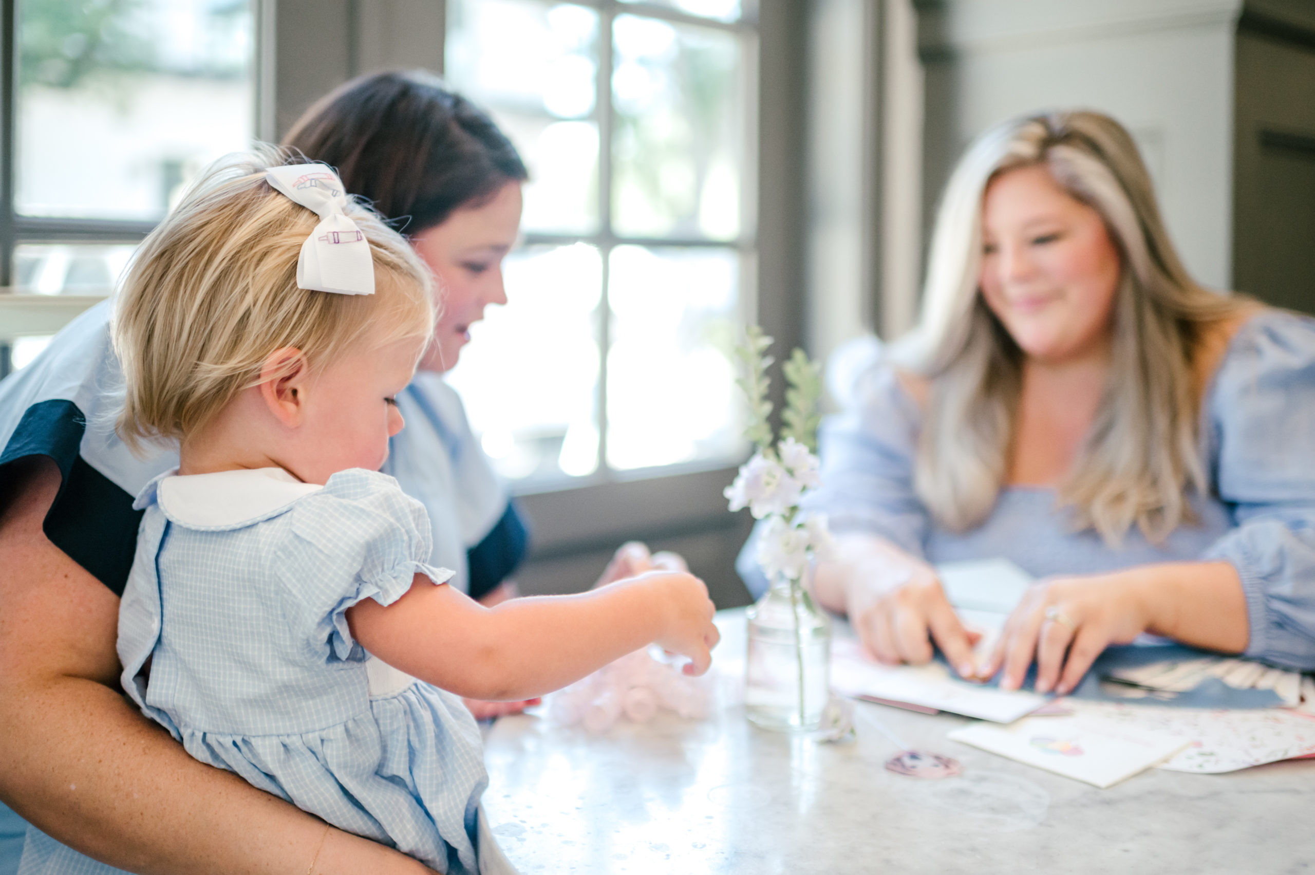 Two woman sitting on table making paper invitations one holding a baby in a blue dress while the other makes paper invitations 