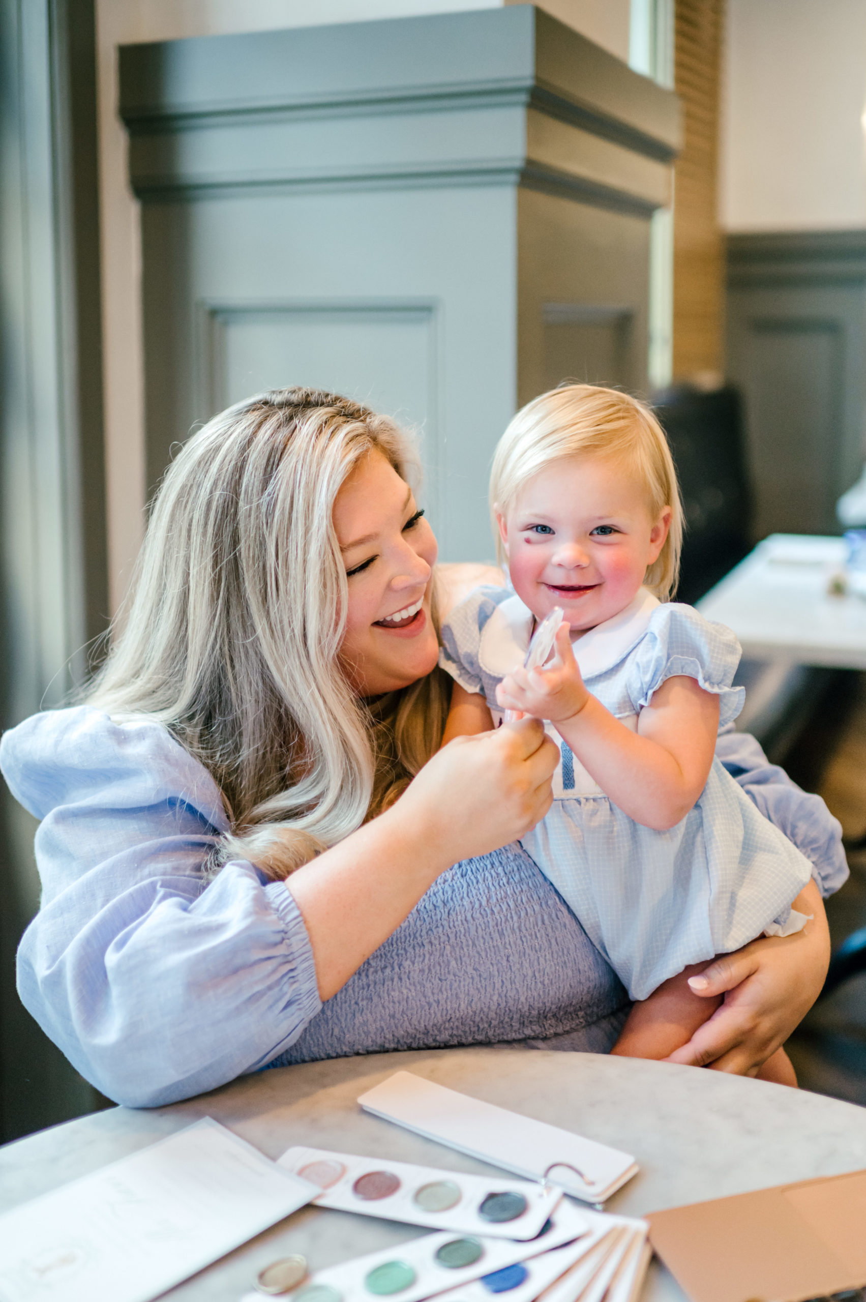 Woman holding baby smiling with paper invitations on table for brand photoshoot