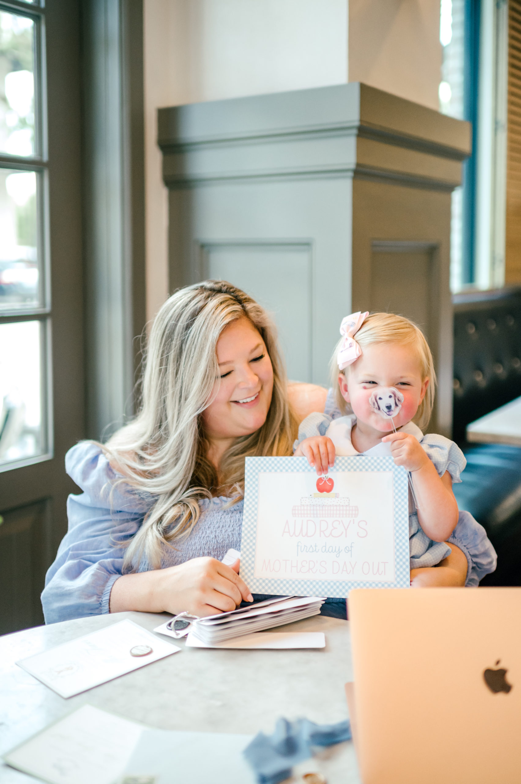 Woman holding baby showing off a milestone card labeled "Audreys first day of mothers day out"