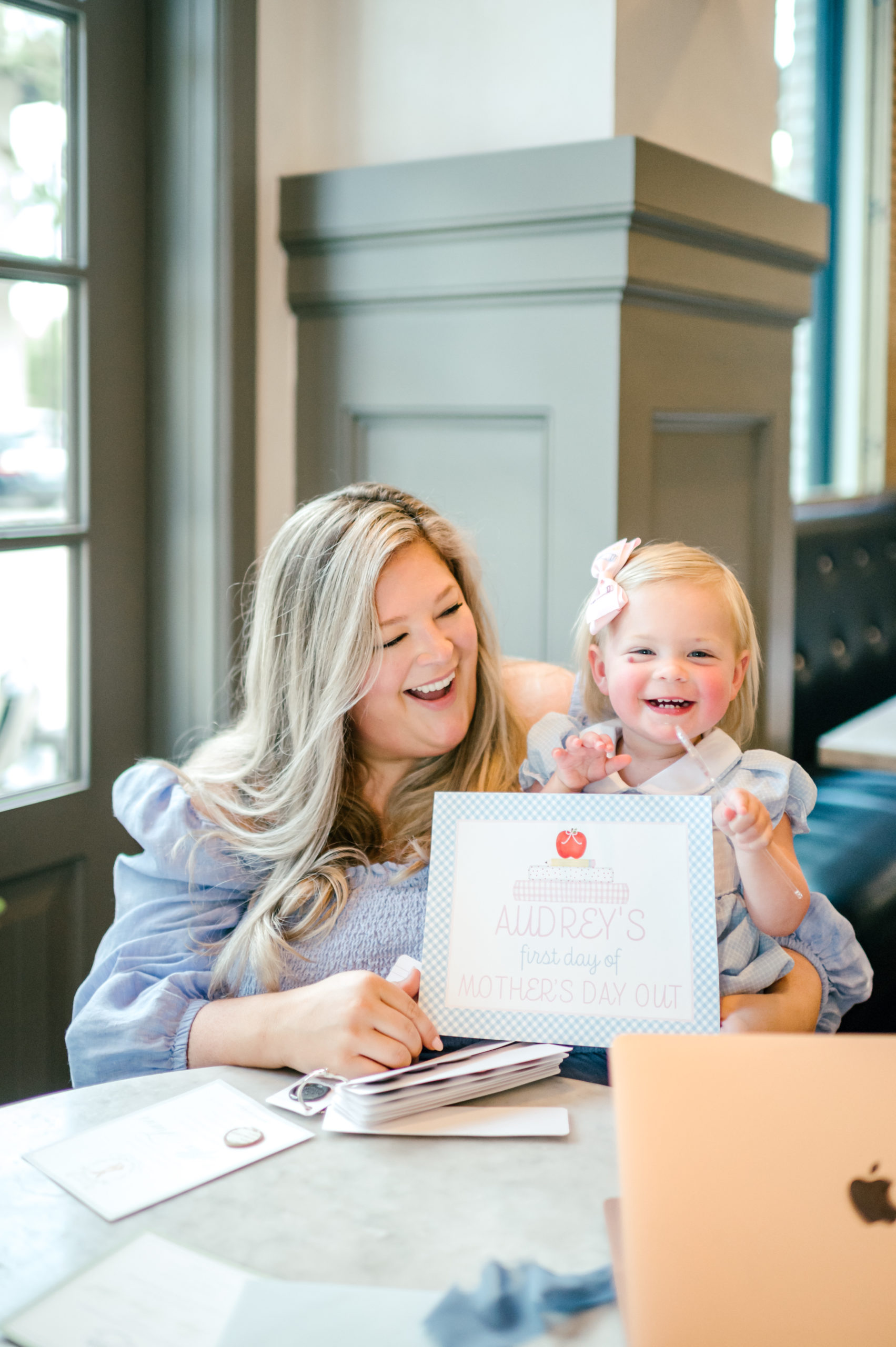 Woman holding baby showing off a milestone card labeled "Audreys first day of mothers day out" for brand photoshoot