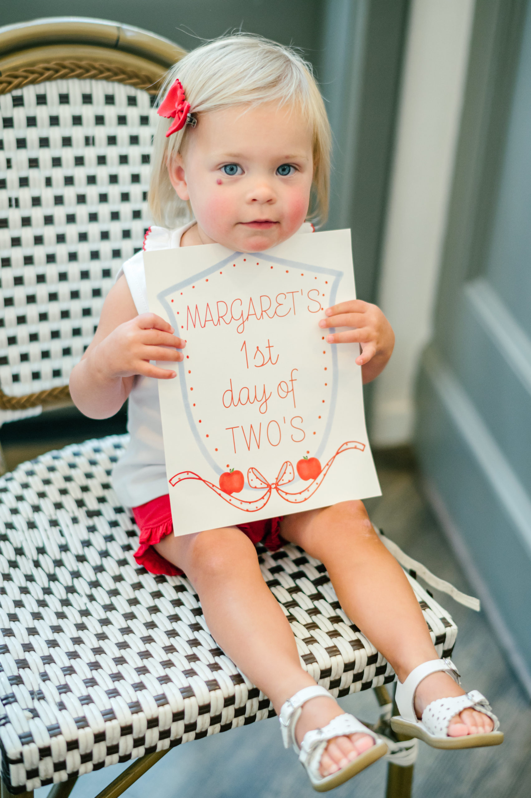 Little girl sitting holding a milestone card labeled "Margaret's 1st day of two's 