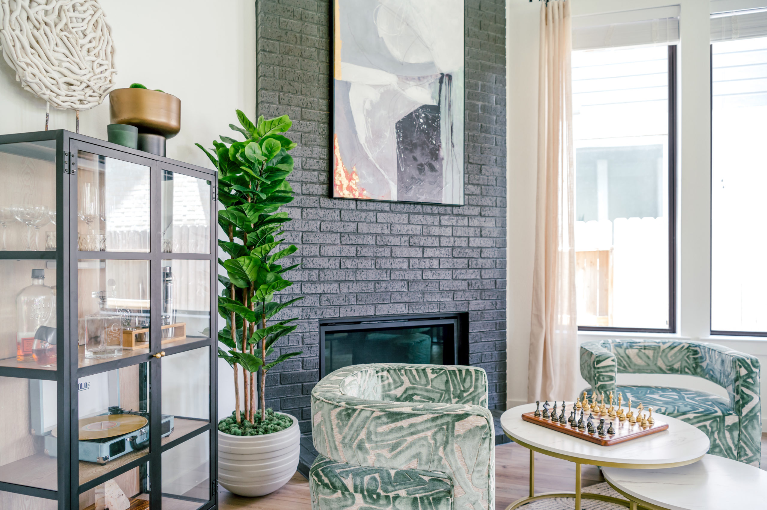 Professional interior photography of living room with a glass cabinet filled of drinkware, alcohol, and a record player, standing next to a green love seat and circle table with chess