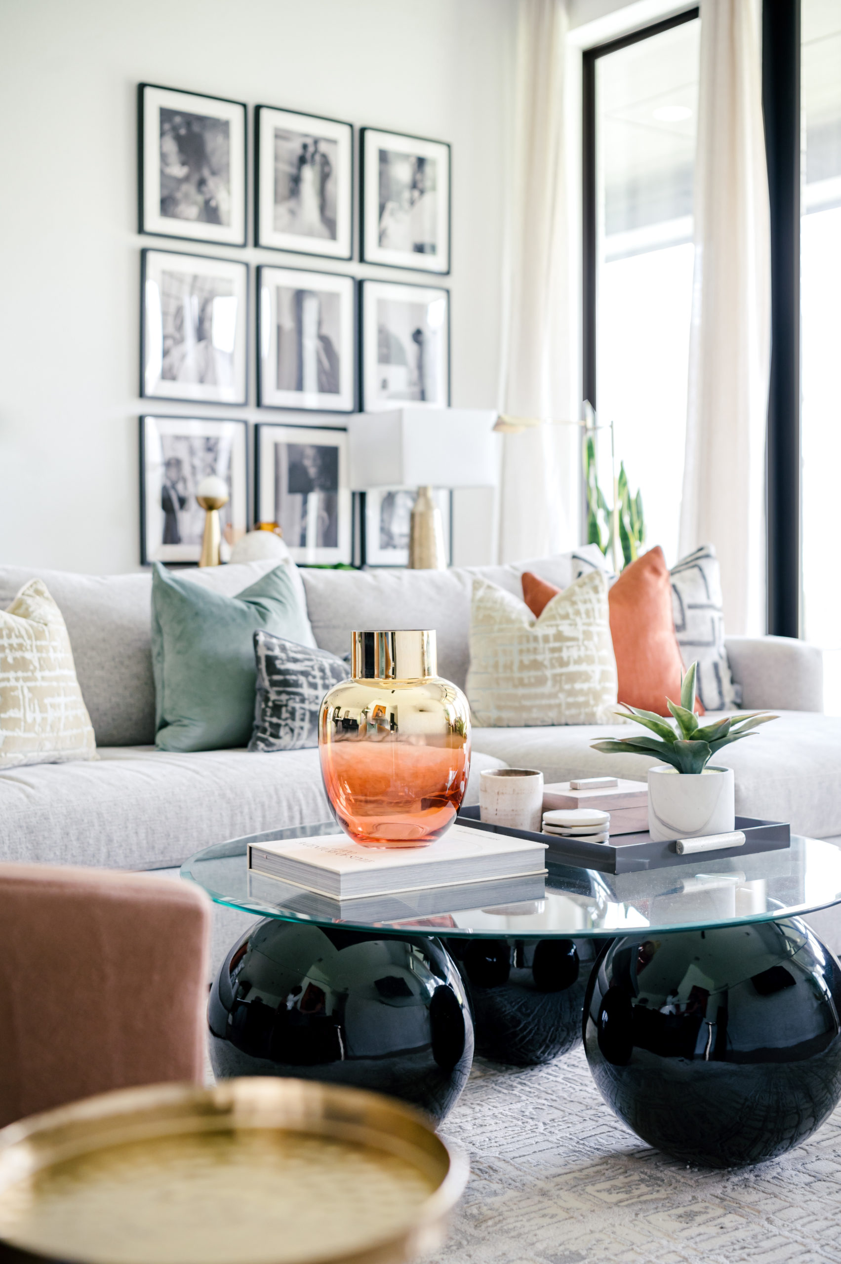 Living room interior design, light grey couch with blue, white, and orange pillows, in front of a wall with family photos hanging, behind a glass table