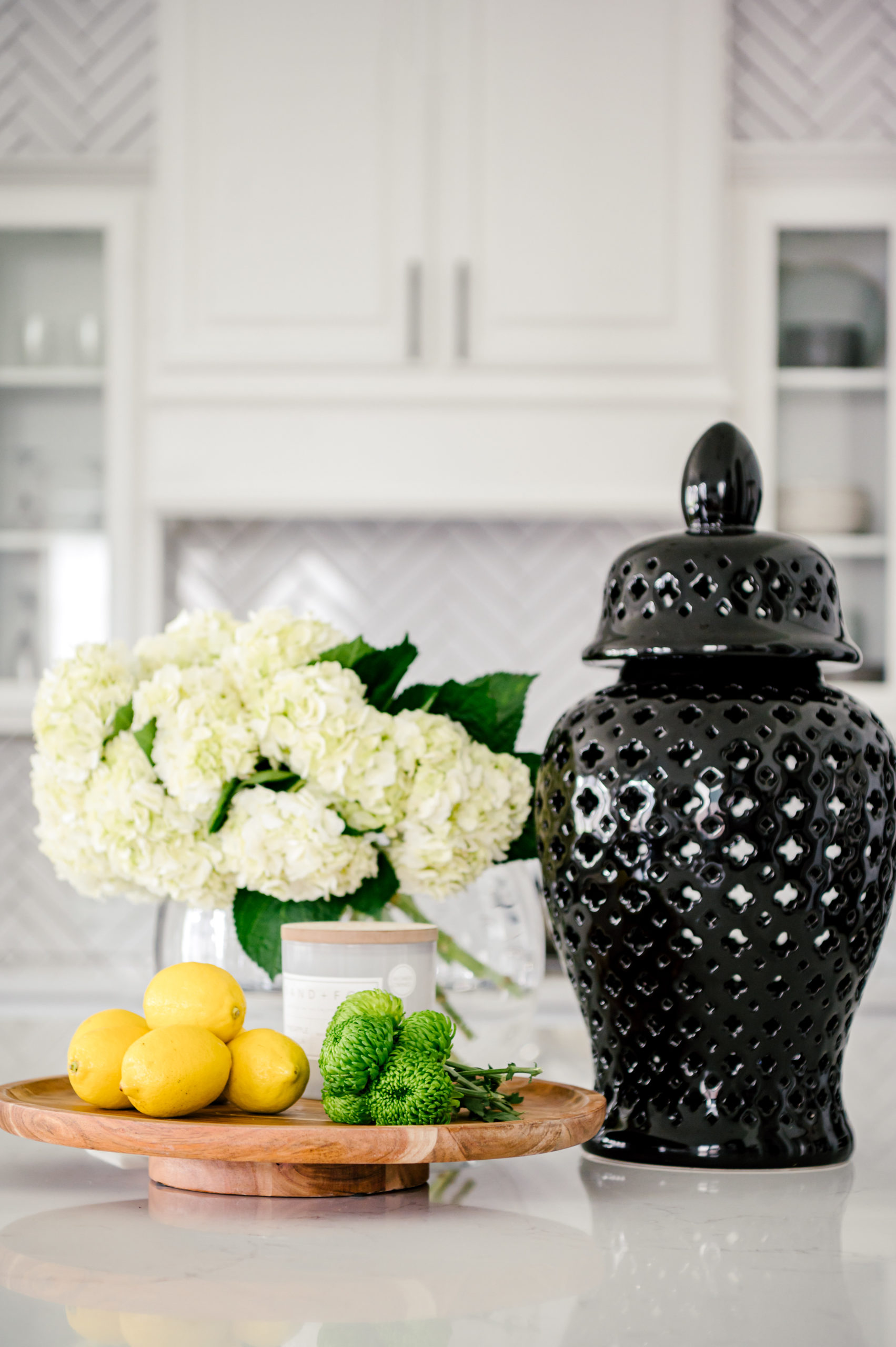 White roses in a glass vase next to a black vase and plate with lemons