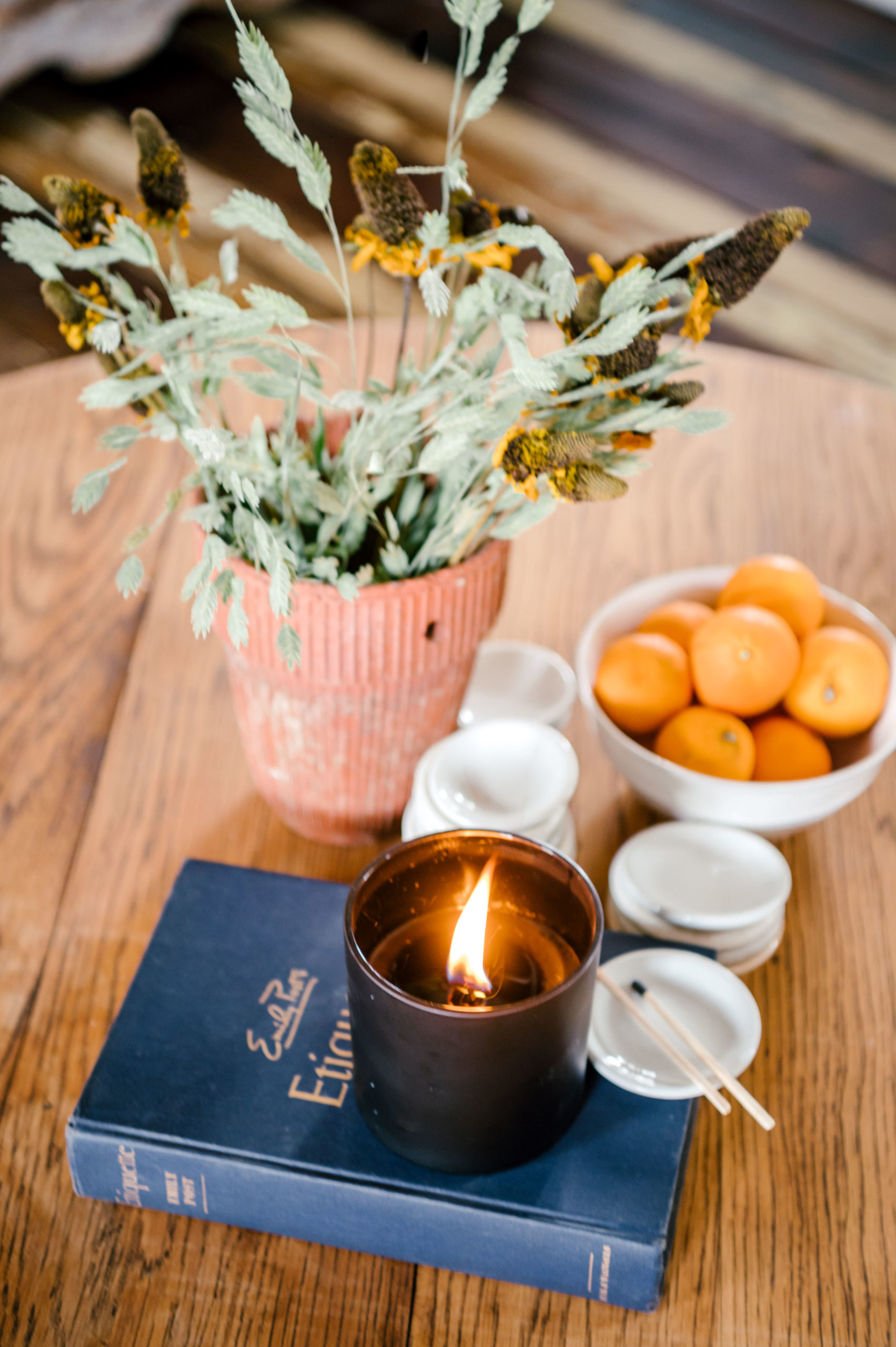 Plant, bowl of oranges, lit candle, and book on a wooden table