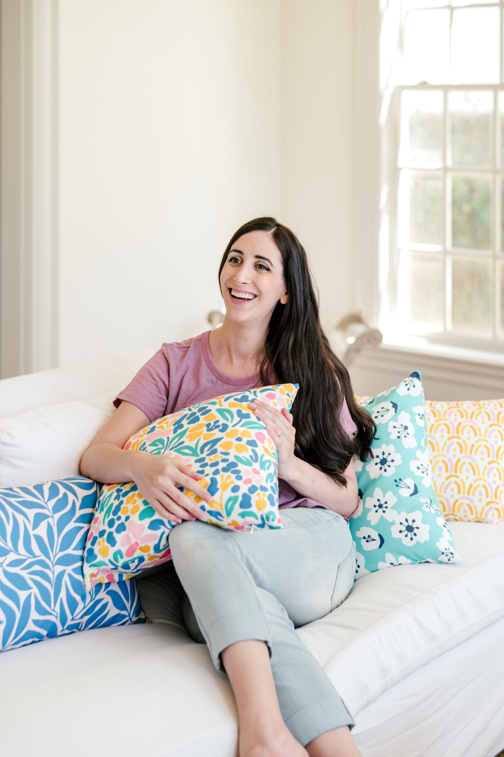Woman designer sitting on a white couch holding colorful pillows that she designed herself smiling for her branding session