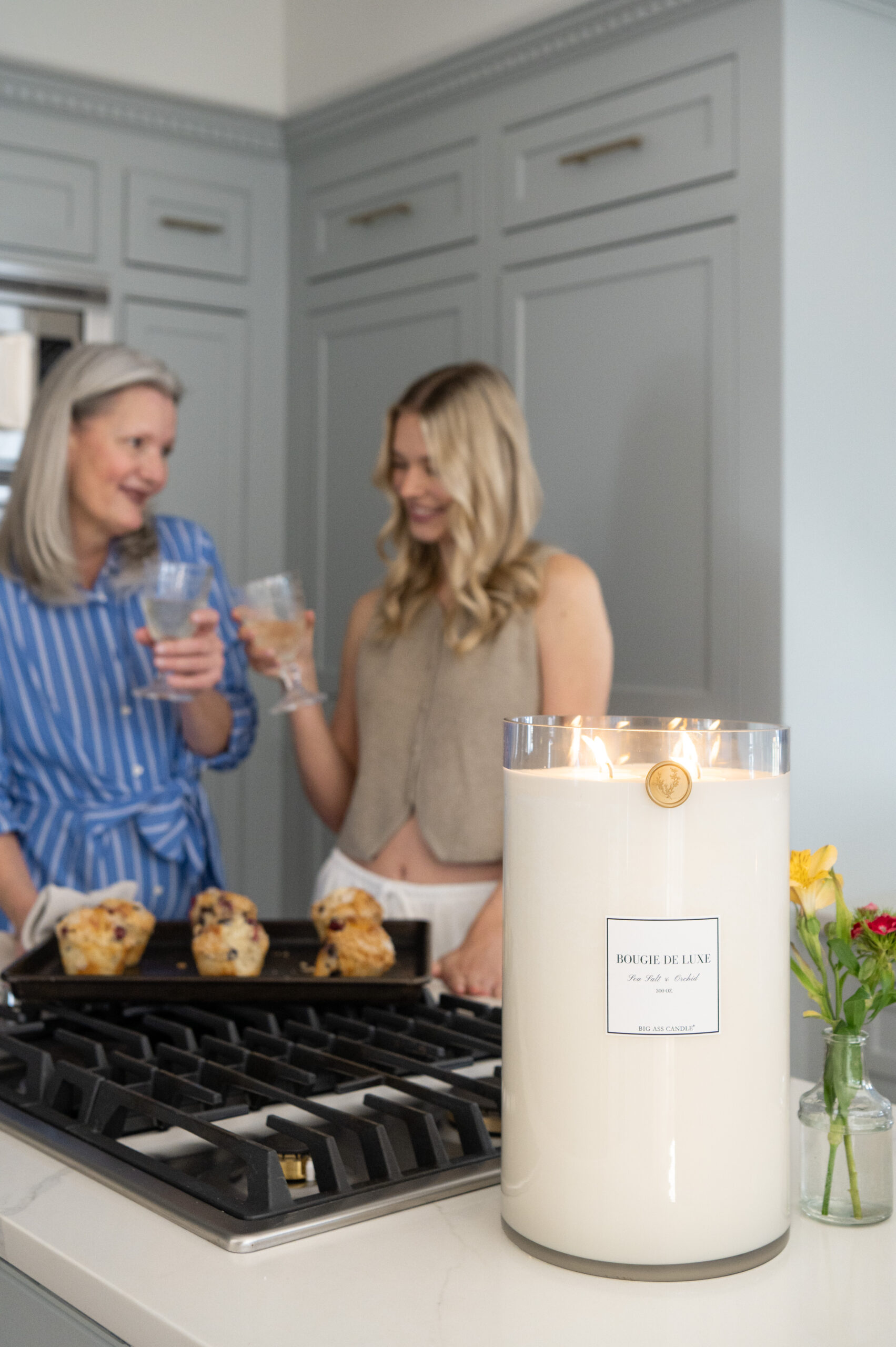 Two women holding glasses of sparkling drinks while standing near a kitchen counter, surrounded by seasonal ingredients and a lit candle—perfect for branding photos capturing joyful moments.