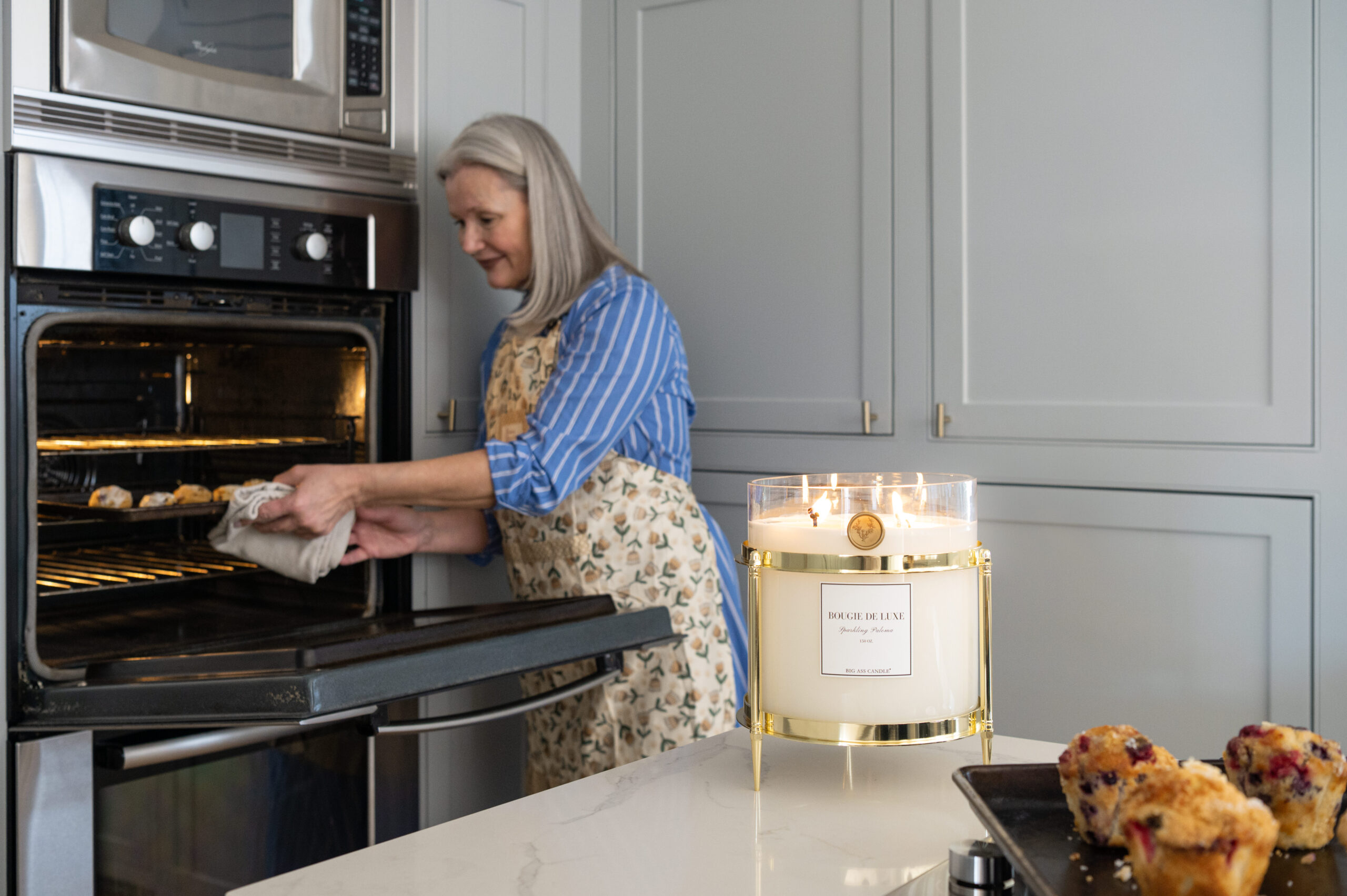 A woman pulling freshly baked muffins out of the oven, with a lit candle beside her, showcasing a cozy home atmosphere