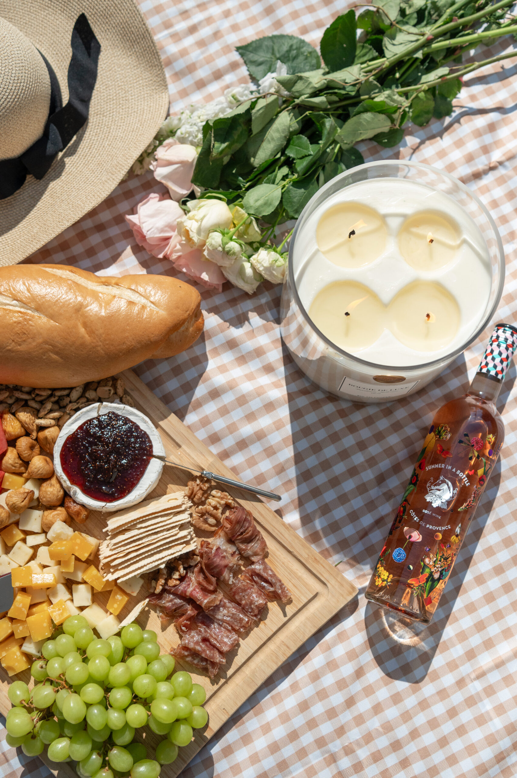 A beautiful arrangement of cheeses, fruits, and a bottle of wine, surrounded by seasonal florals and a candle, photographed in a vibrant and warm summer setting.