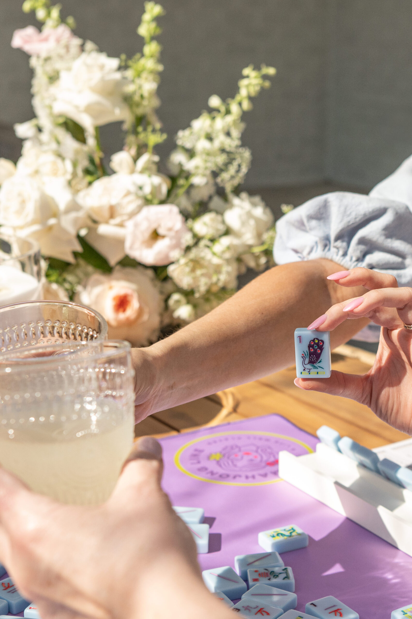 A close-up of a hand holding a Mahjong tile and a drink, with a stunning floral arrangement in the background, perfect for showcasing seasonal photography in a casual, vibrant setting.