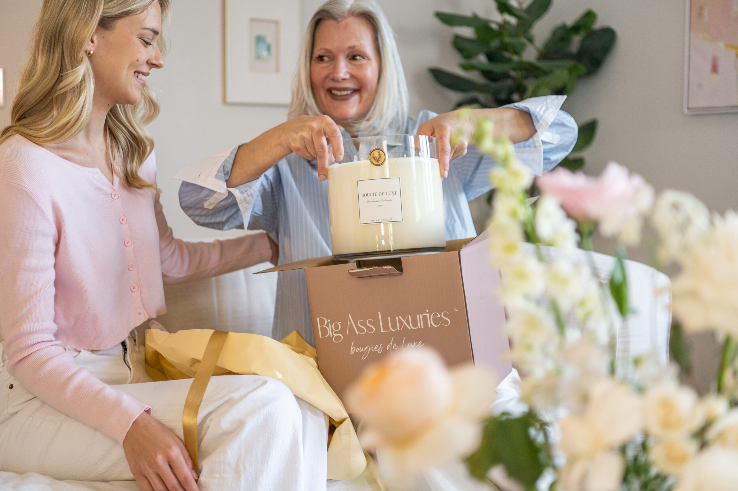 A woman receiving a gift box containing luxury candles, surrounded by delicate flowers and soft lighting, captured in a moment of warmth and excitement perfect for seasonal branding photography.

